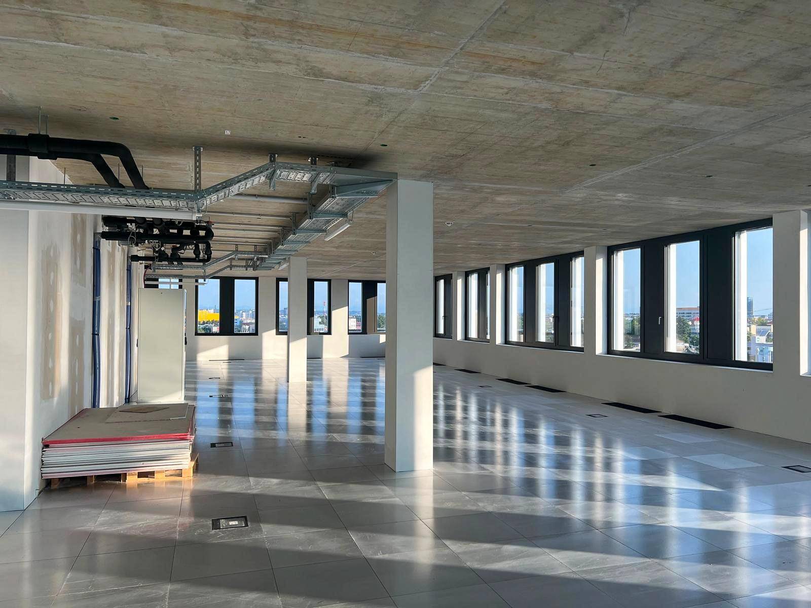 Unausgebauter Bürobereich mit hohen Fenstern, Betondecke und sichtbaren Installationen.