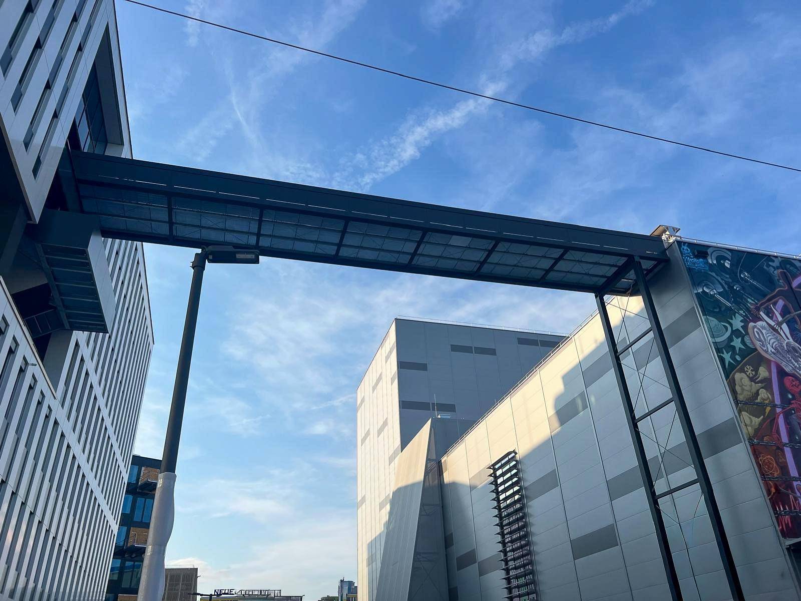 Eine moderne Fußgängerbrücke verbindet zwei Gebäudeteile unter einem strahlend blauen Himmel.