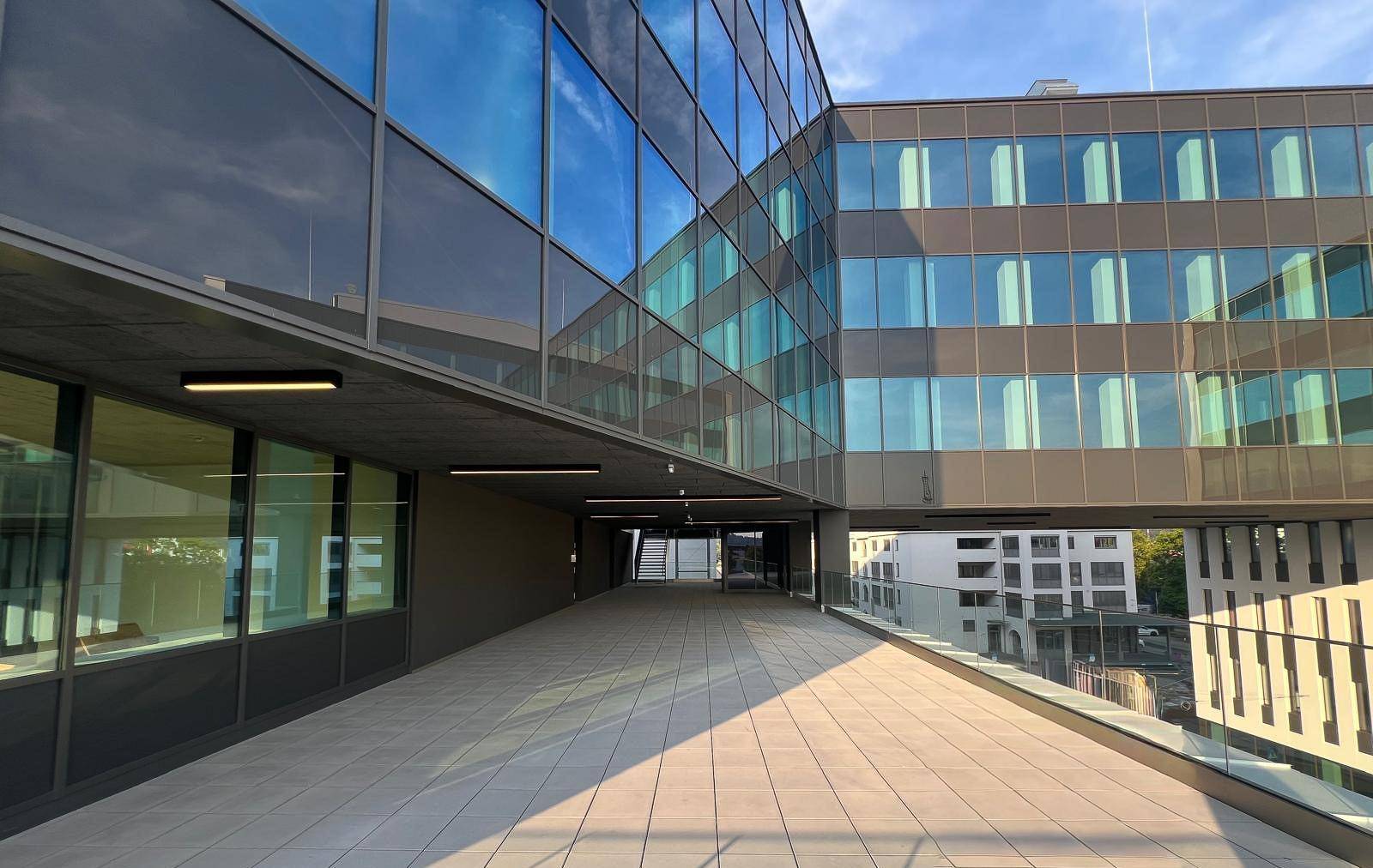 Eine weitläufige Terrasse mit Glasgeländer und Blick auf die moderne Gebäudefassade und den Himmel.