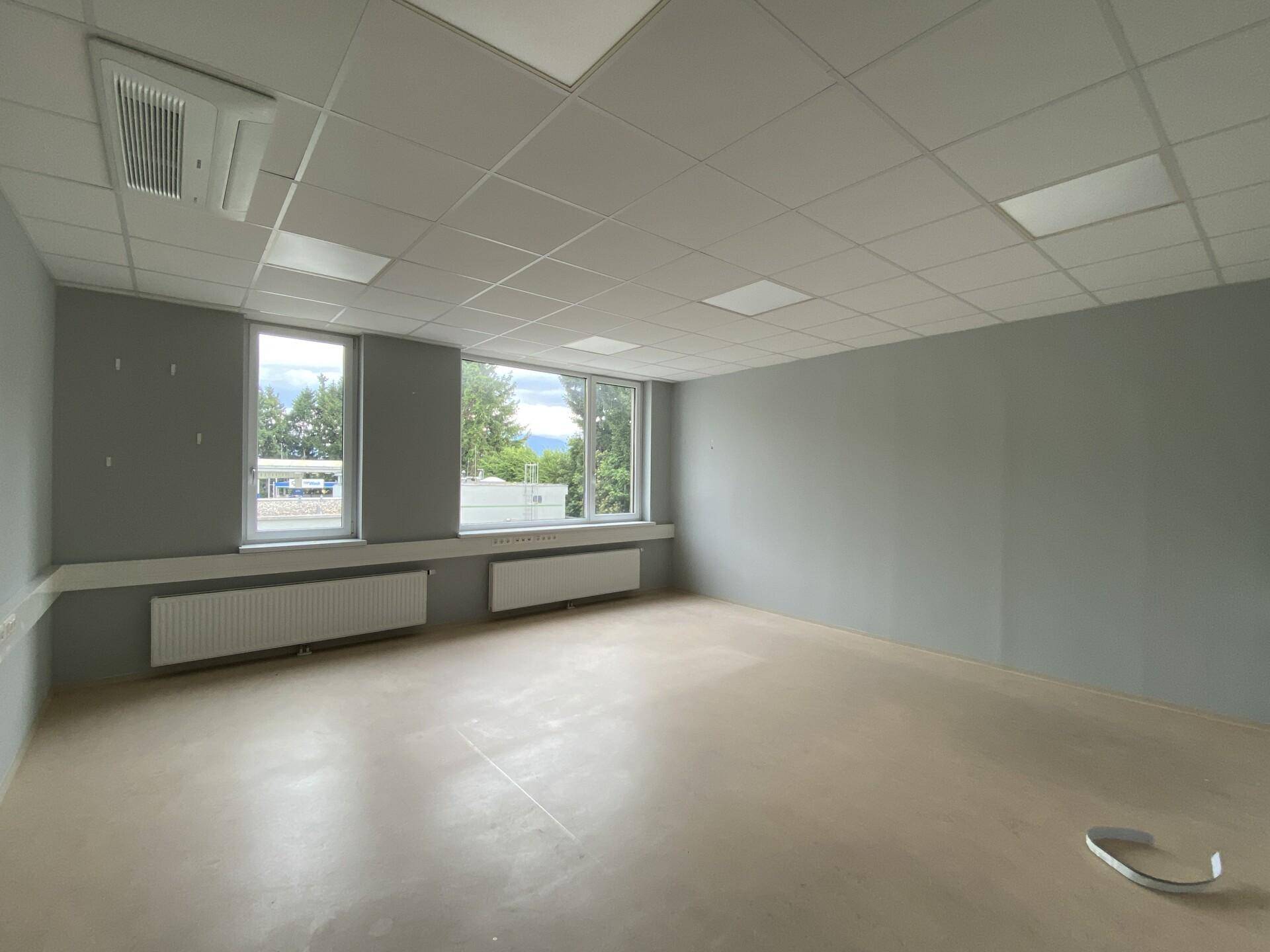 Leerer Büroraum mit hellgrauen Wänden, beigem Boden und Blick auf die Umgebung durch die Fenster.