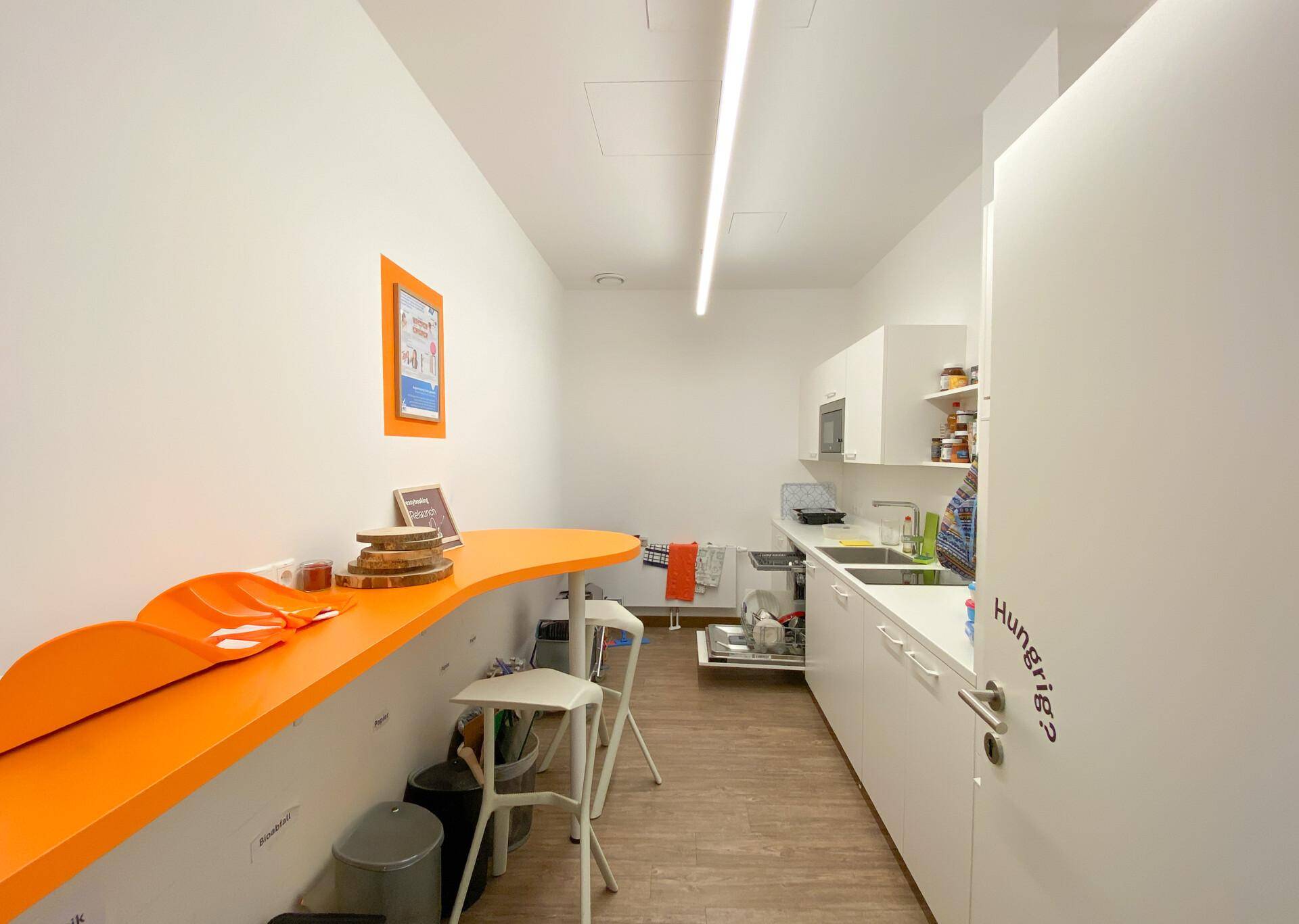 Geräumige Küche oder Pausenraum mit weißen Schränken, einer langen orangefarbenen Theke und Spülmaschine.