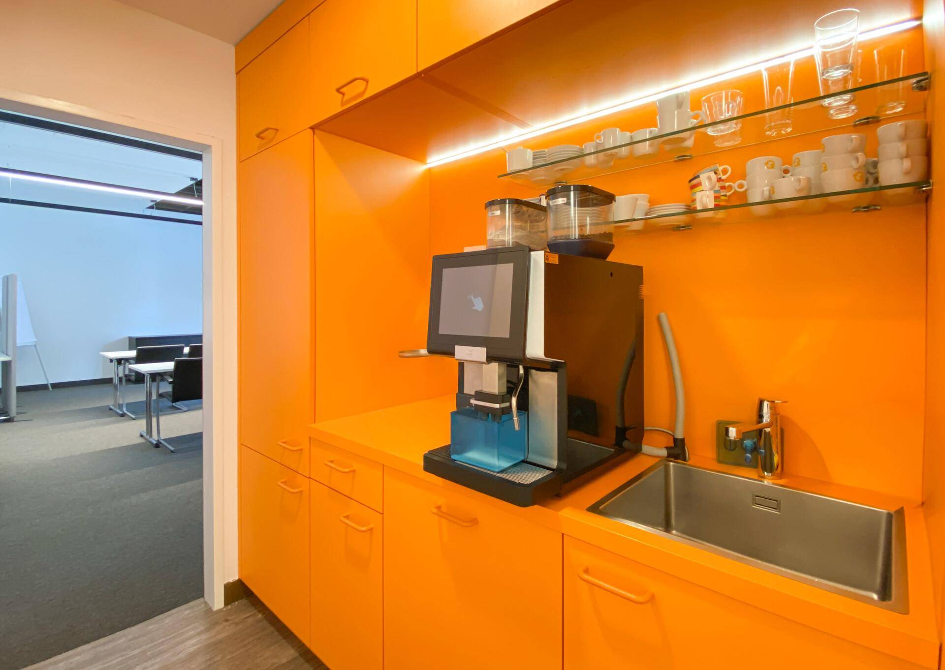 Moderne Teeküche mit leuchtend orangefarbenen Schränken, integrierter Kaffeemaschine und Spülbecken.