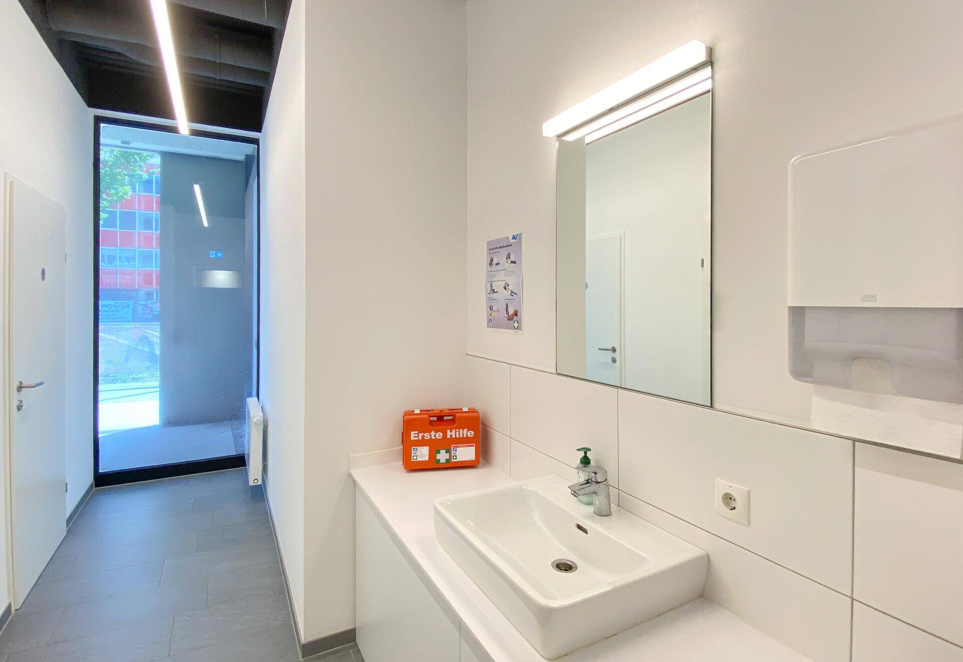 Modernes Badezimmer mit weißem Waschbecken, großem Spiegel und hellen Fliesen, gut beleuchtet.