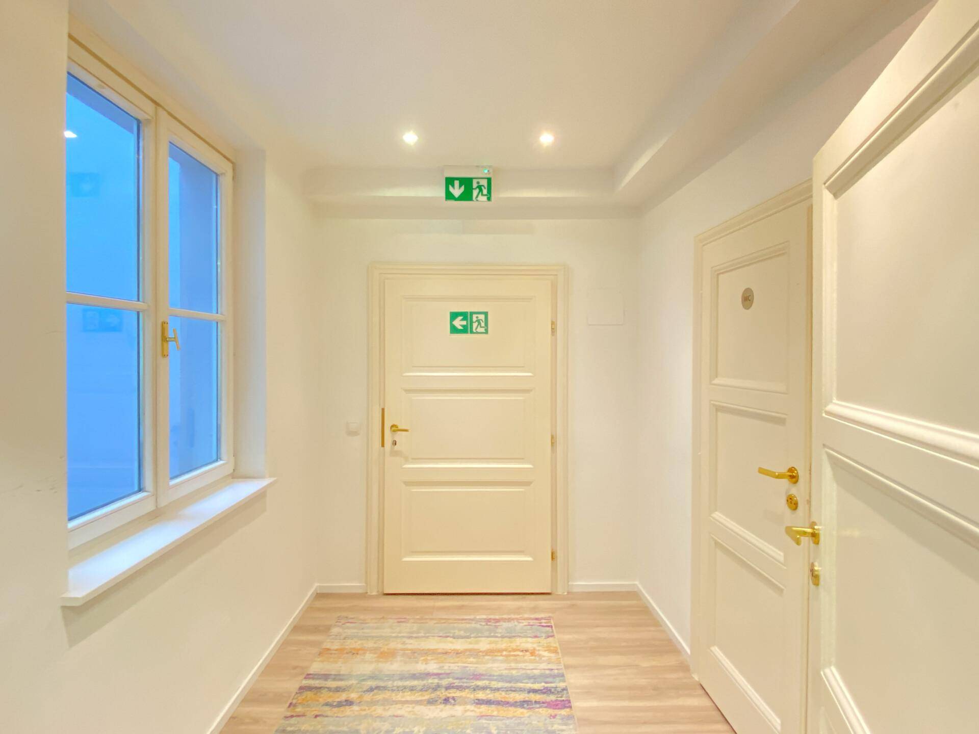 Flur mit mehreren Türen, einem Fenster und deutlichen Notausgangsschildern.