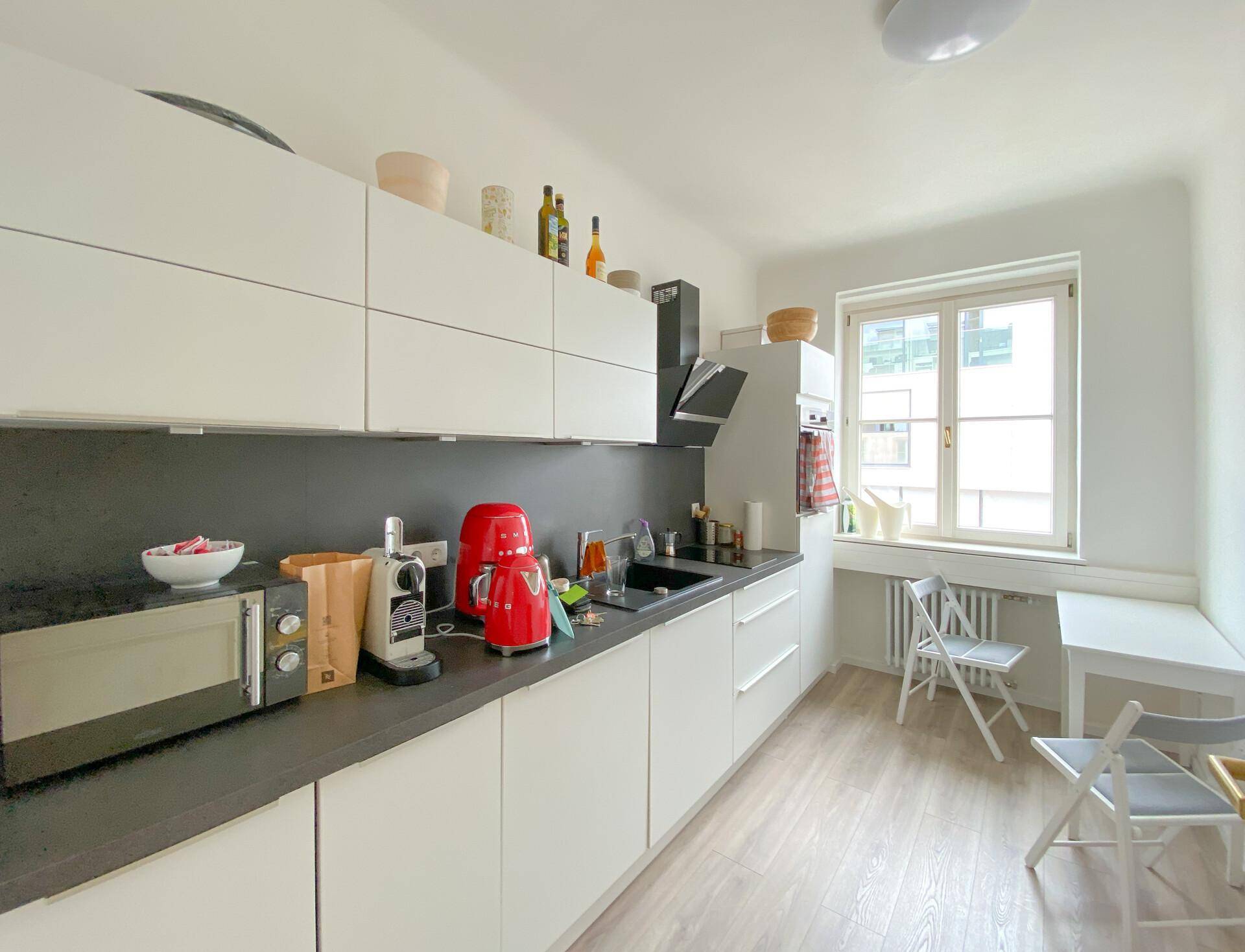 Moderne Küche mit weißen Schränken, dunklen Arbeitsplatten und einem kleinen Essbereich am Fenster.
