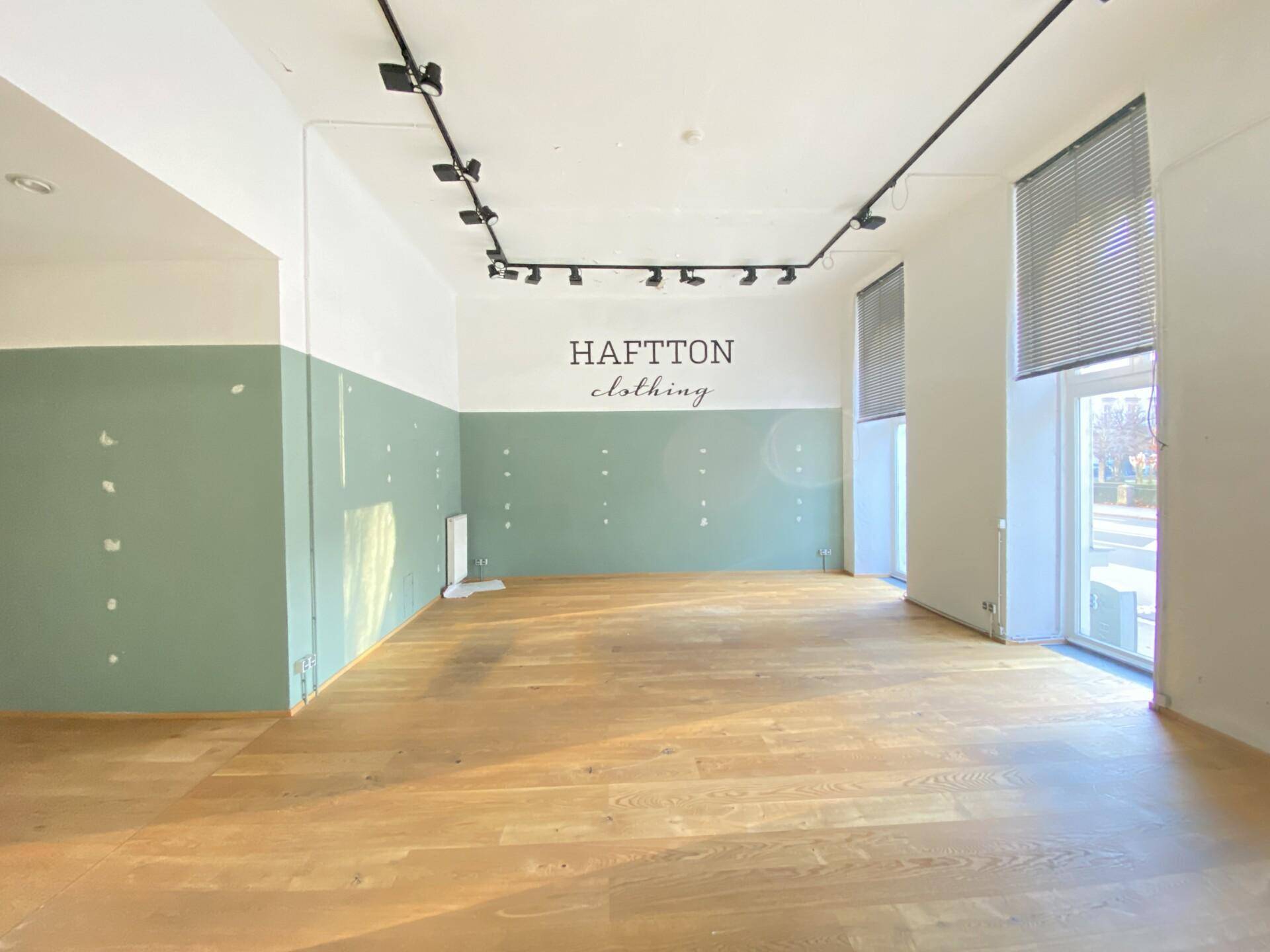Leerer Ladenbereich mit Holzboden, großen Fenstern und einer Wand mit Markenbeschriftung.