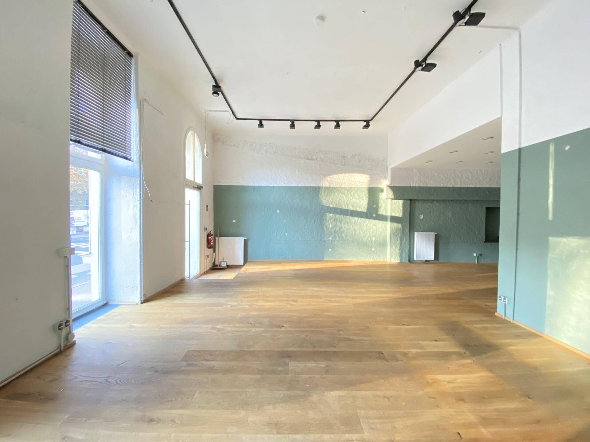 Geräumiger Geschäftsraum mit Holzboden, hohen Decken und viel Tageslicht durch große Fenster.