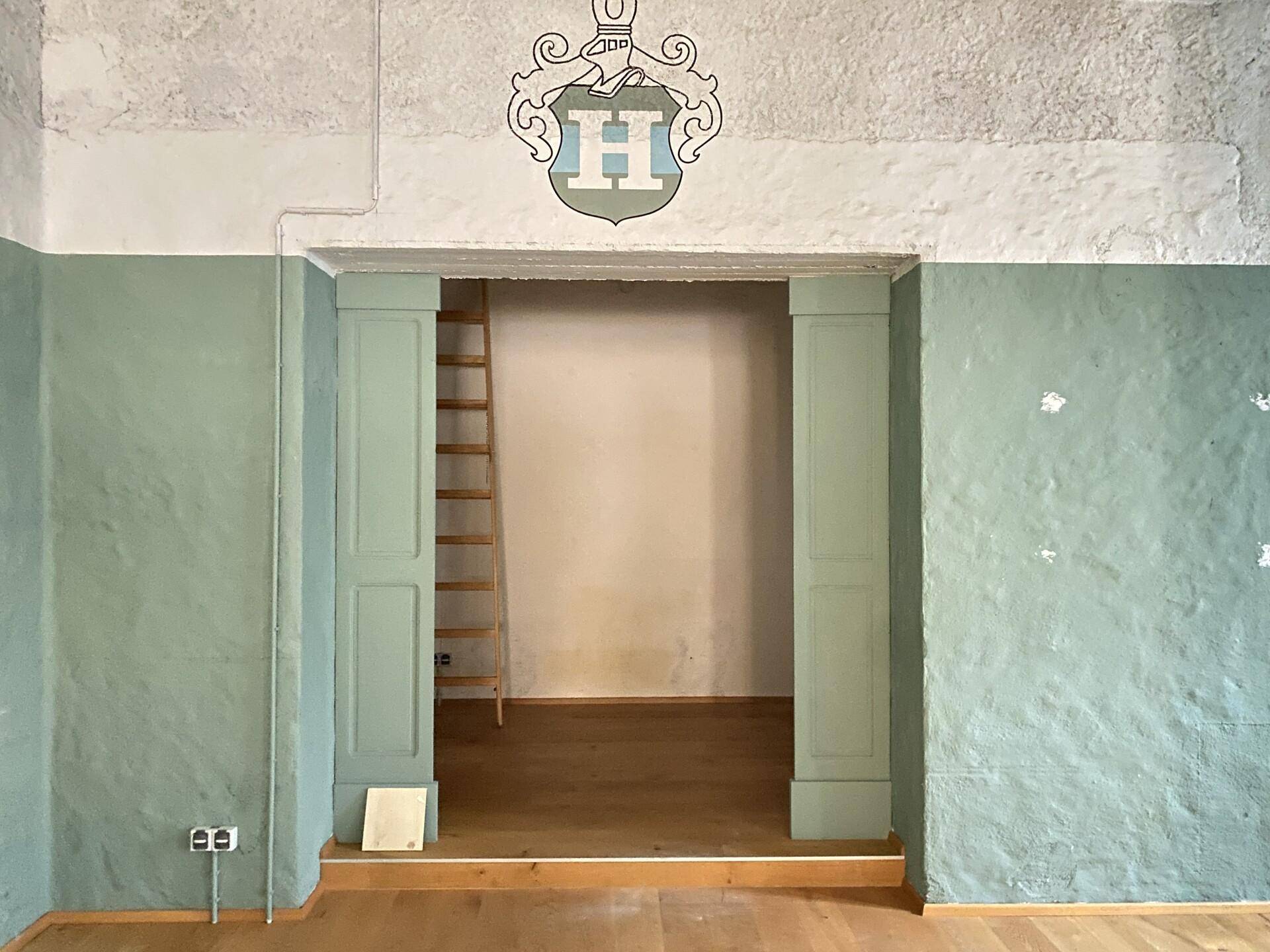 Nischenbereich mit Holzboden, einer Leiter und einem Wappen an der Wand.