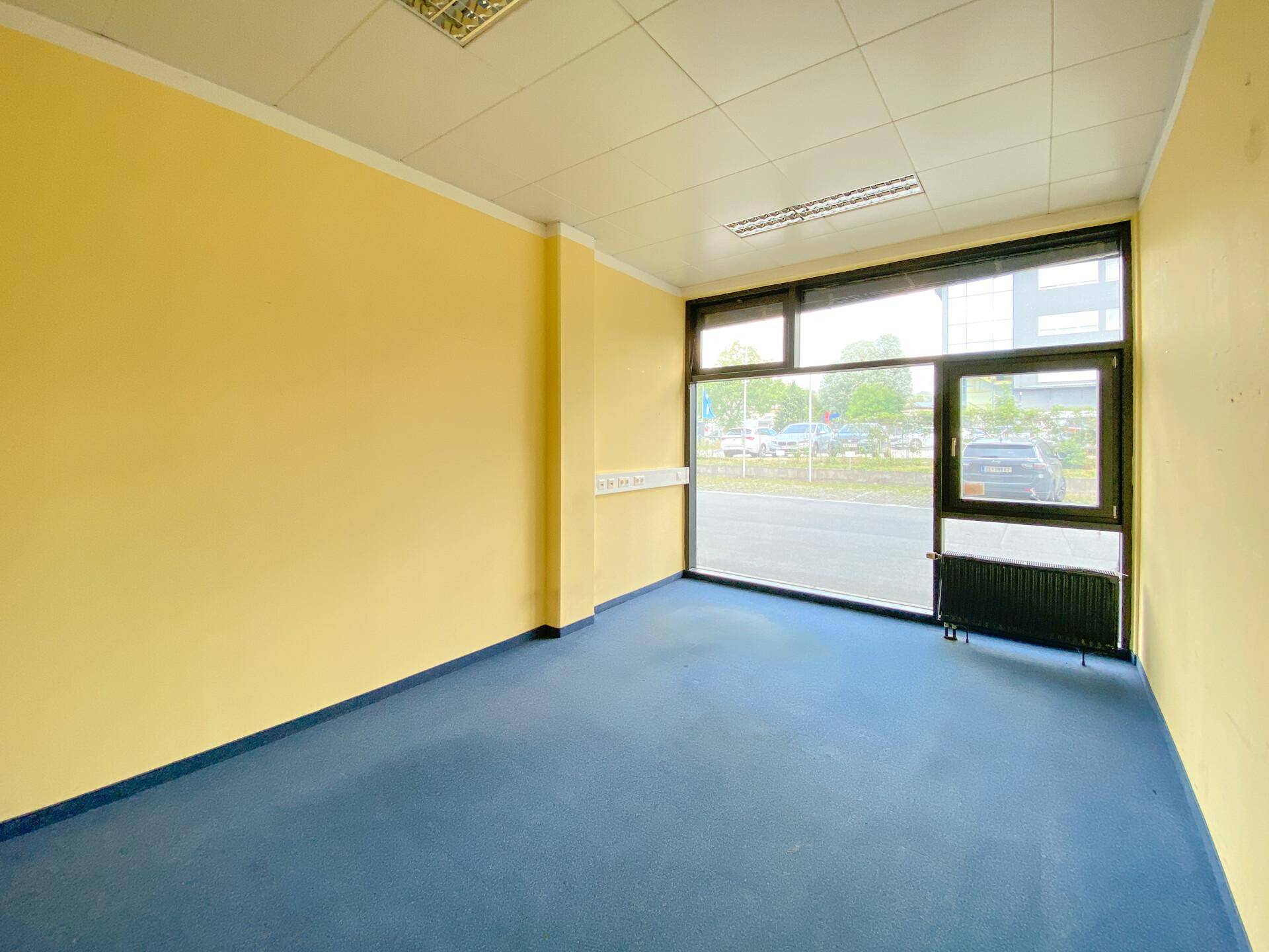 Helles Büro mit gelben Wänden, blauem Teppichboden und großen Fenstern zur Straße.