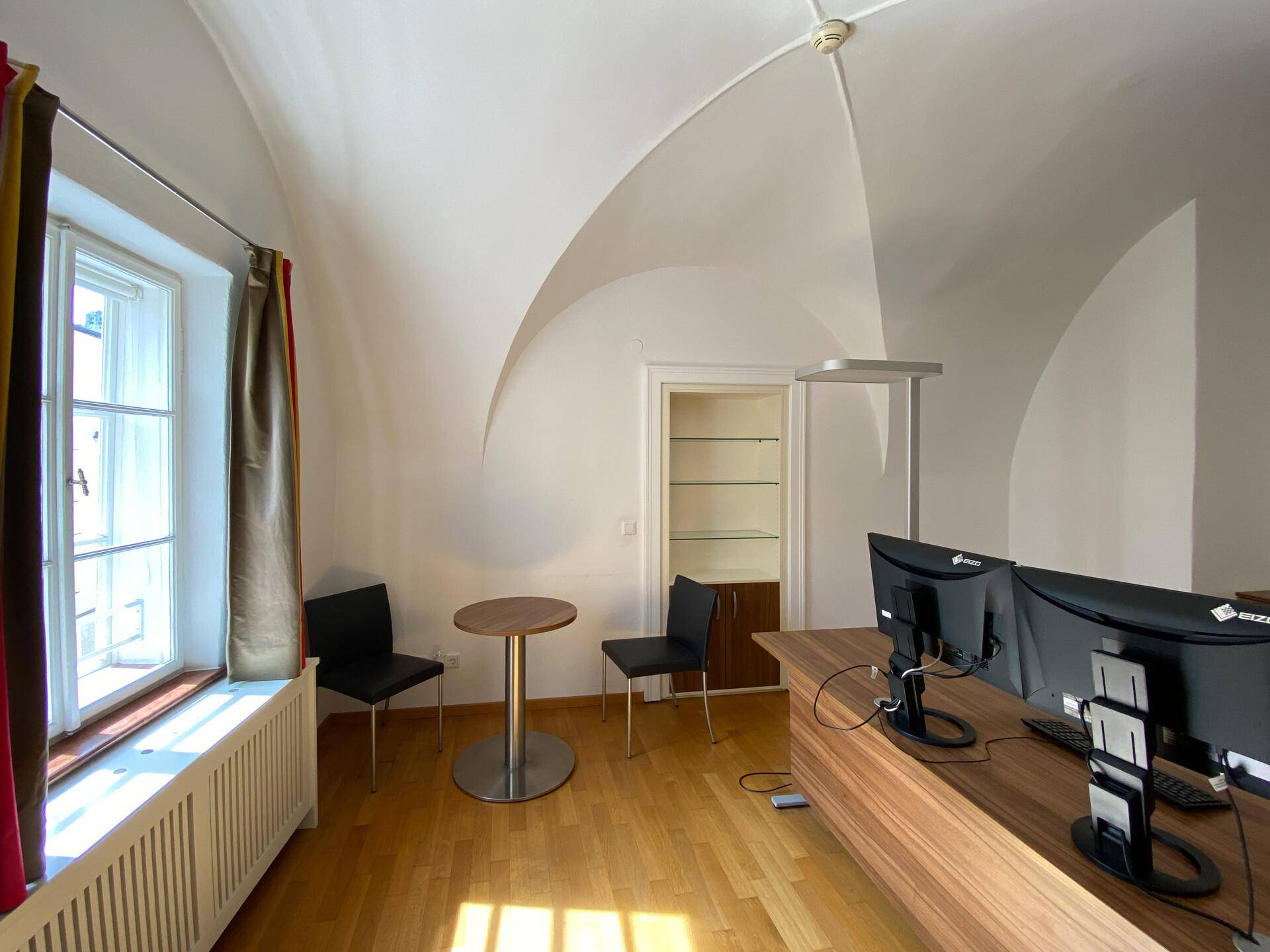 Helles Büro mit Gewölbedecke, großem Fenster und einem modernen Arbeitsplatz mit Sitzgelegenheiten.