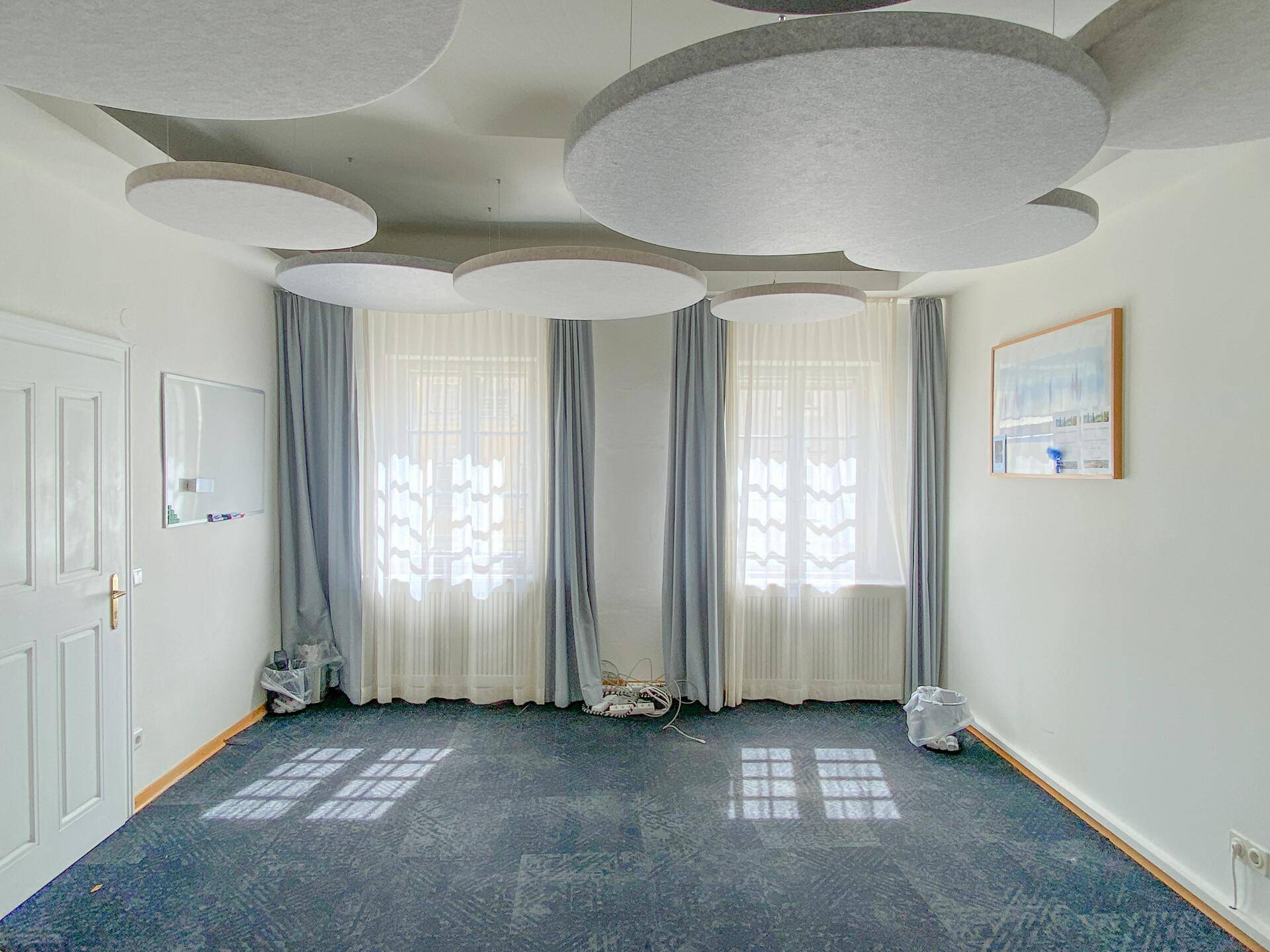 Leerer Büroraum mit dunklem Teppichboden, großen Fenstern und schallabsorbierenden Deckenpaneelen.