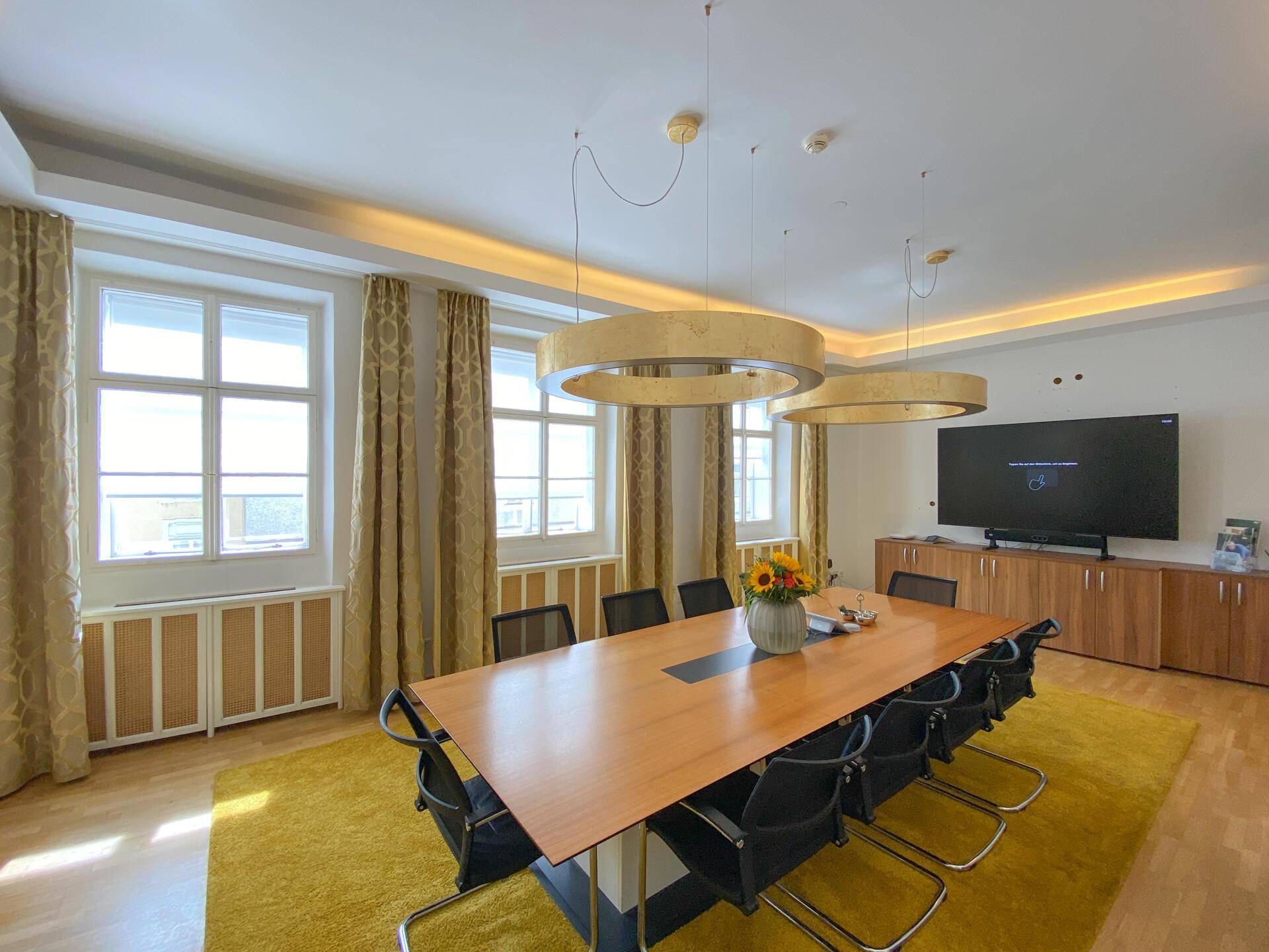 Geräumiger Konferenzraum mit großem Bildschirm, Holzboden und stilvollen Vorhängen an den hohen Fenstern.