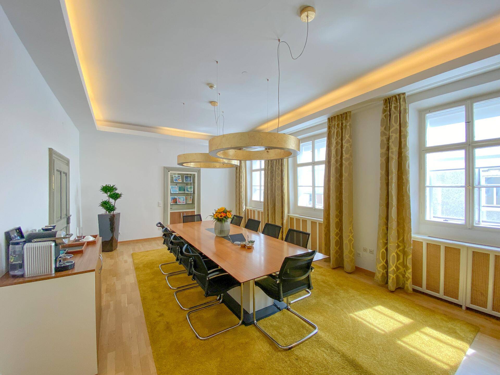 Großer Konferenzraum mit Holzboden, gelbem Teppich und eleganten goldenen Hängeleuchten über dem Tisch.