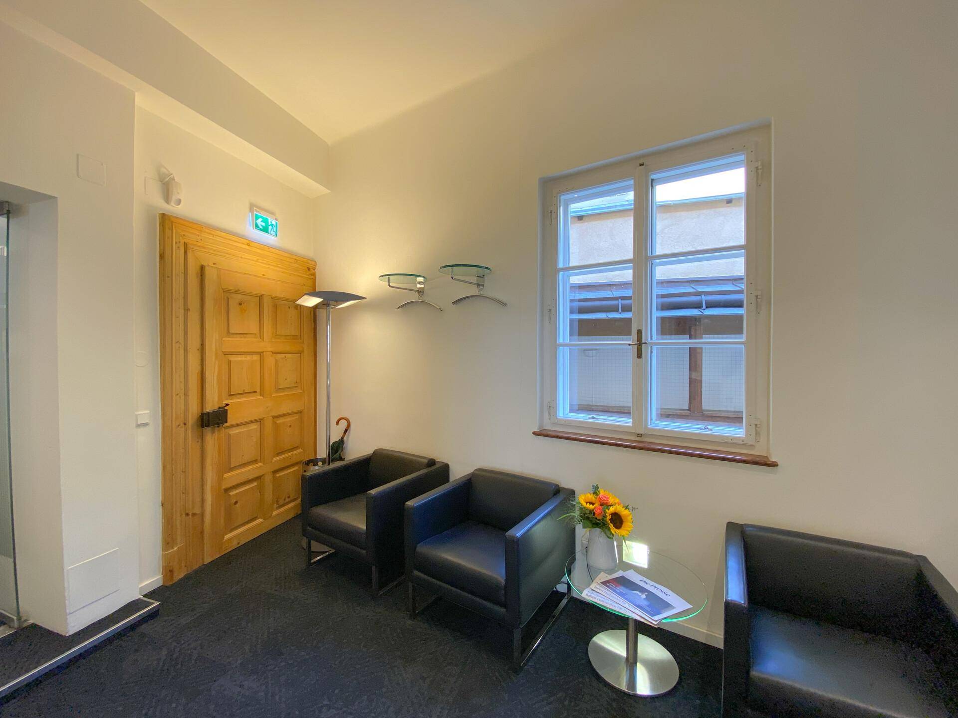 Gemütlicher Wartebereich mit Ledersesseln, einem kleinen Tisch und einem Fenster mit Blick auf einen Innenhof.