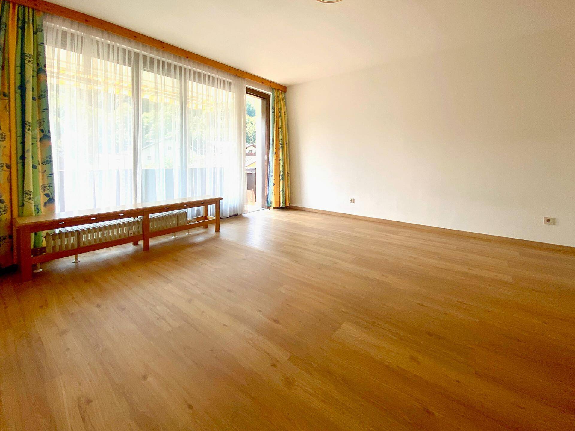 Geräumiges Wohnzimmer mit Holzboden, weißen Wänden und Zugang zu einem Balkon oder Garten.