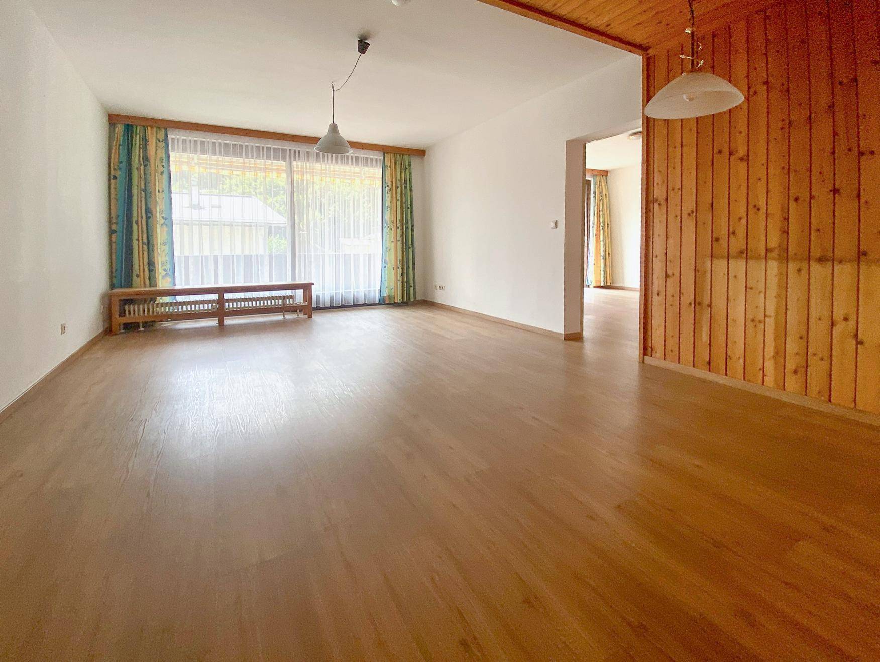 Helles Wohnzimmer mit Holzpaneelen an der Wand, Holzboden und großem Fenster mit Vorhängen.