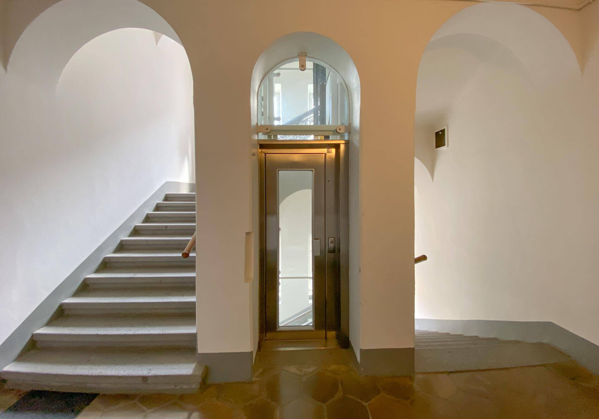 Treppenhaus mit einem modernen Aufzug in der Mitte, umgeben von gewölbten Decken und Treppenaufgängen.
