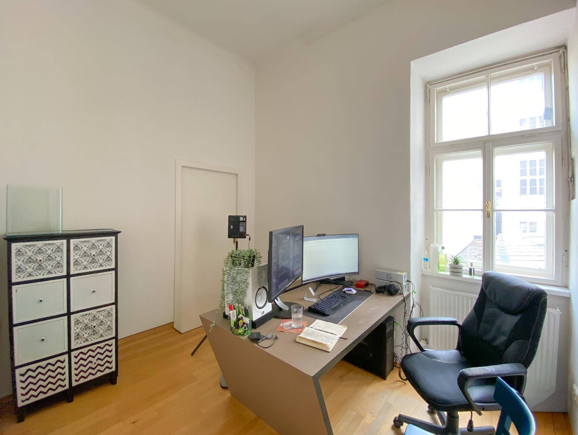 Kleineres Büro mit einem einzelnen Arbeitsplatz, einem dekorativen Schrank und einem großen Fenster für Tageslicht.