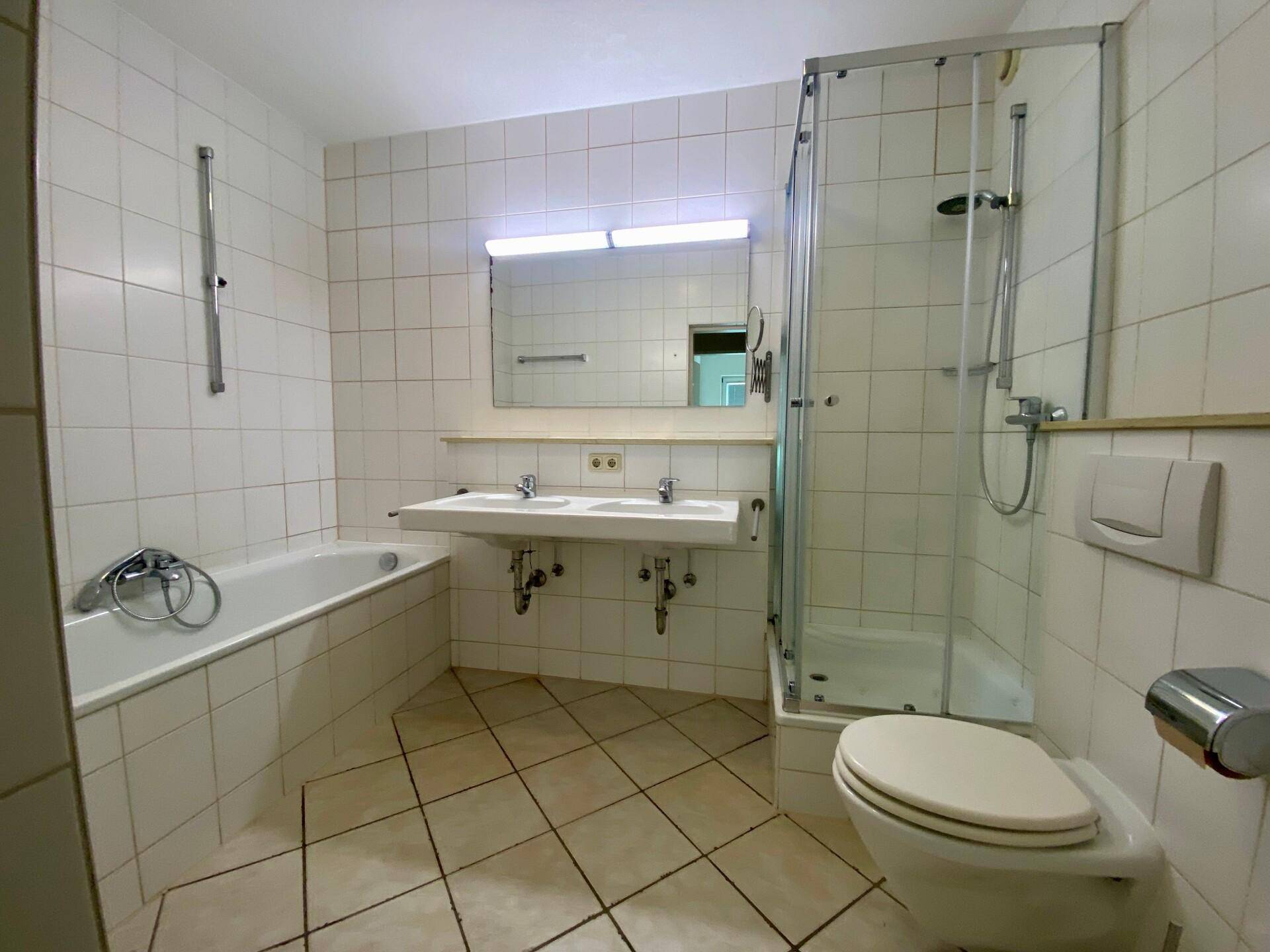 Badezimmer mit weißen Fliesen, Badewanne, Duschkabine, Toilette und Doppelwaschbecken.