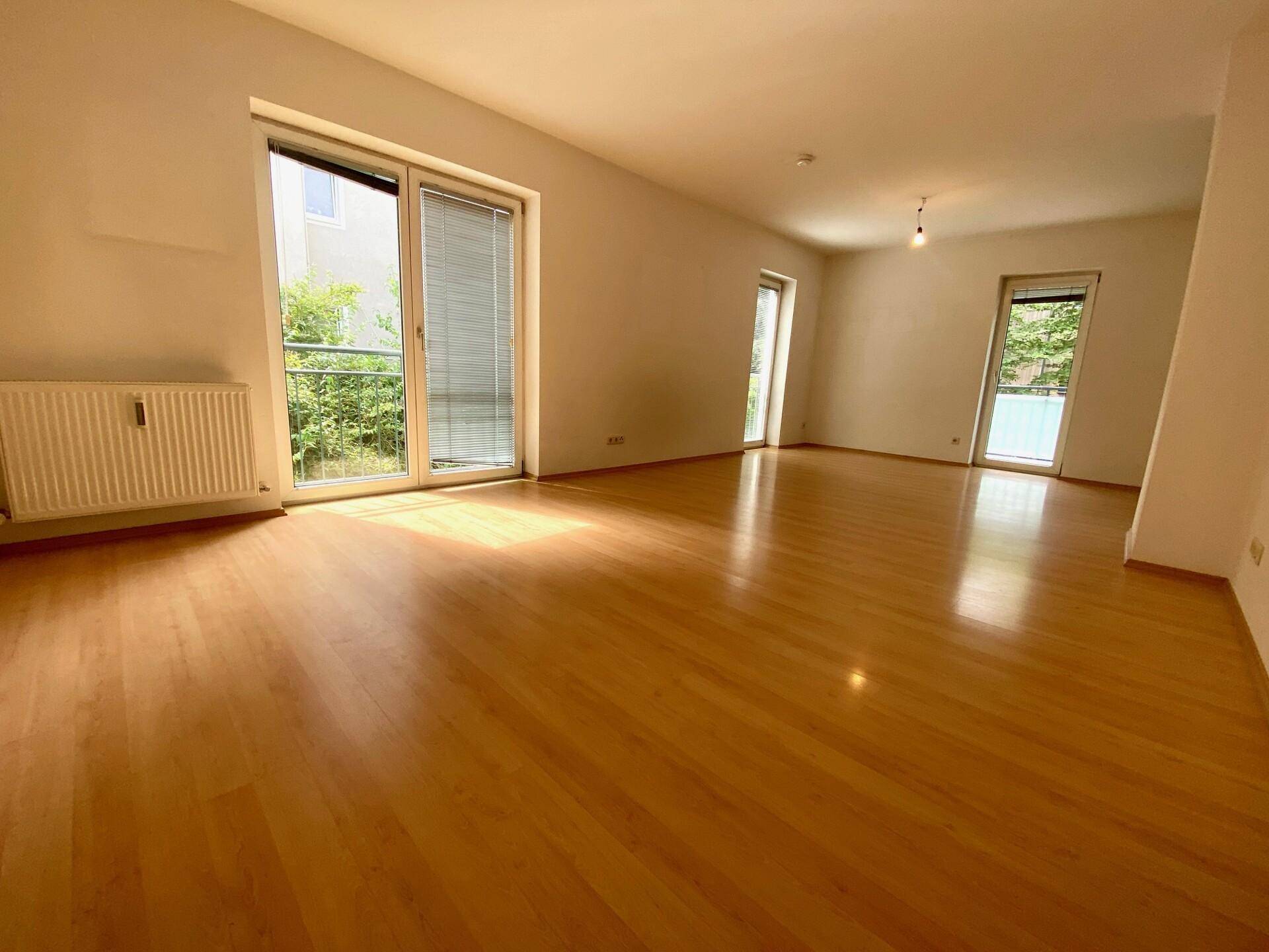 Heller, leerer Wohnraum mit hellem Holzfußboden und großen Fenstern, die viel Tageslicht hereinlassen.