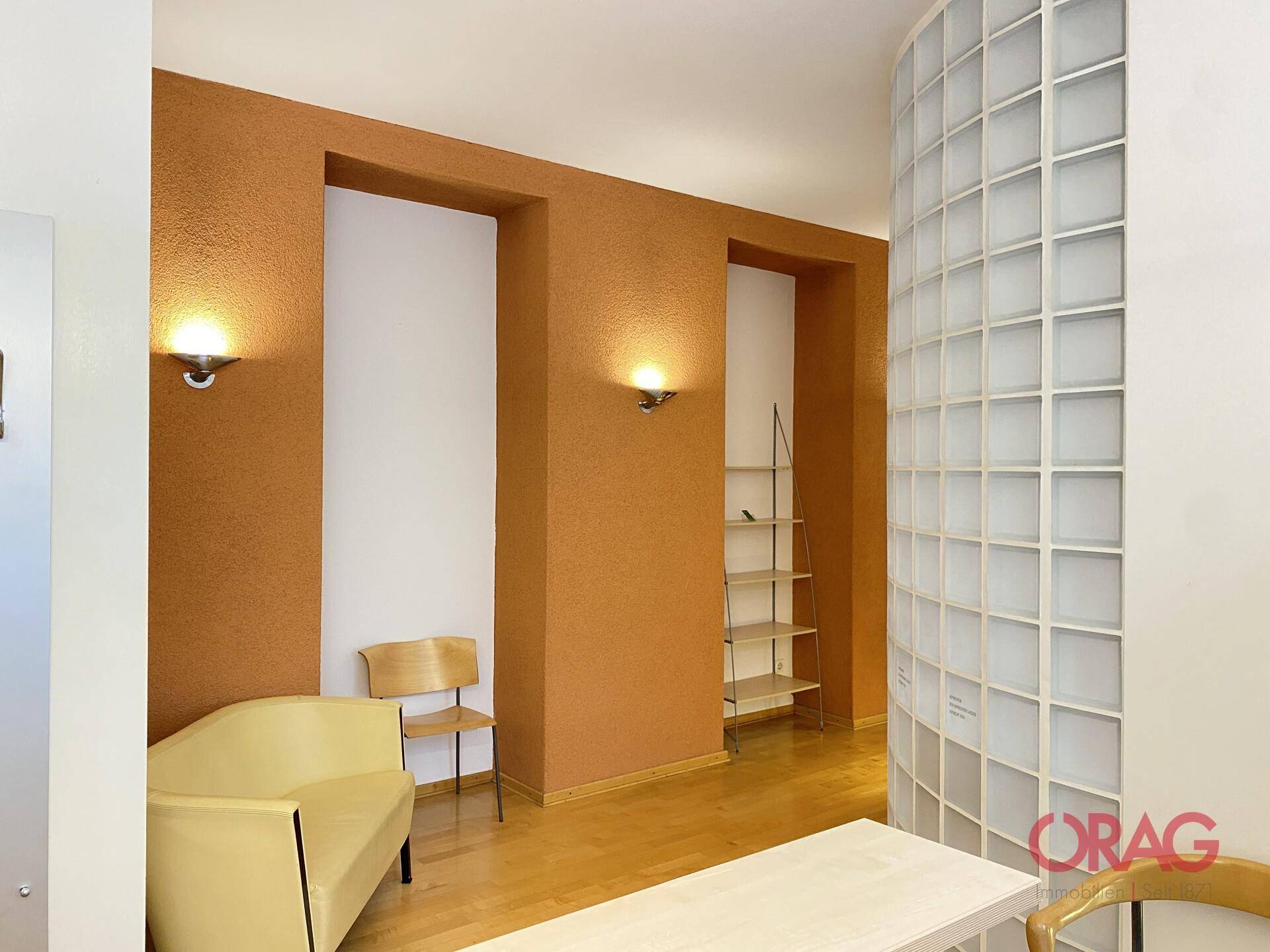 Wartebereich mit orangefarbener Strukturwand, beiger Sitzbank und geschwungener Glasbausteinwand.