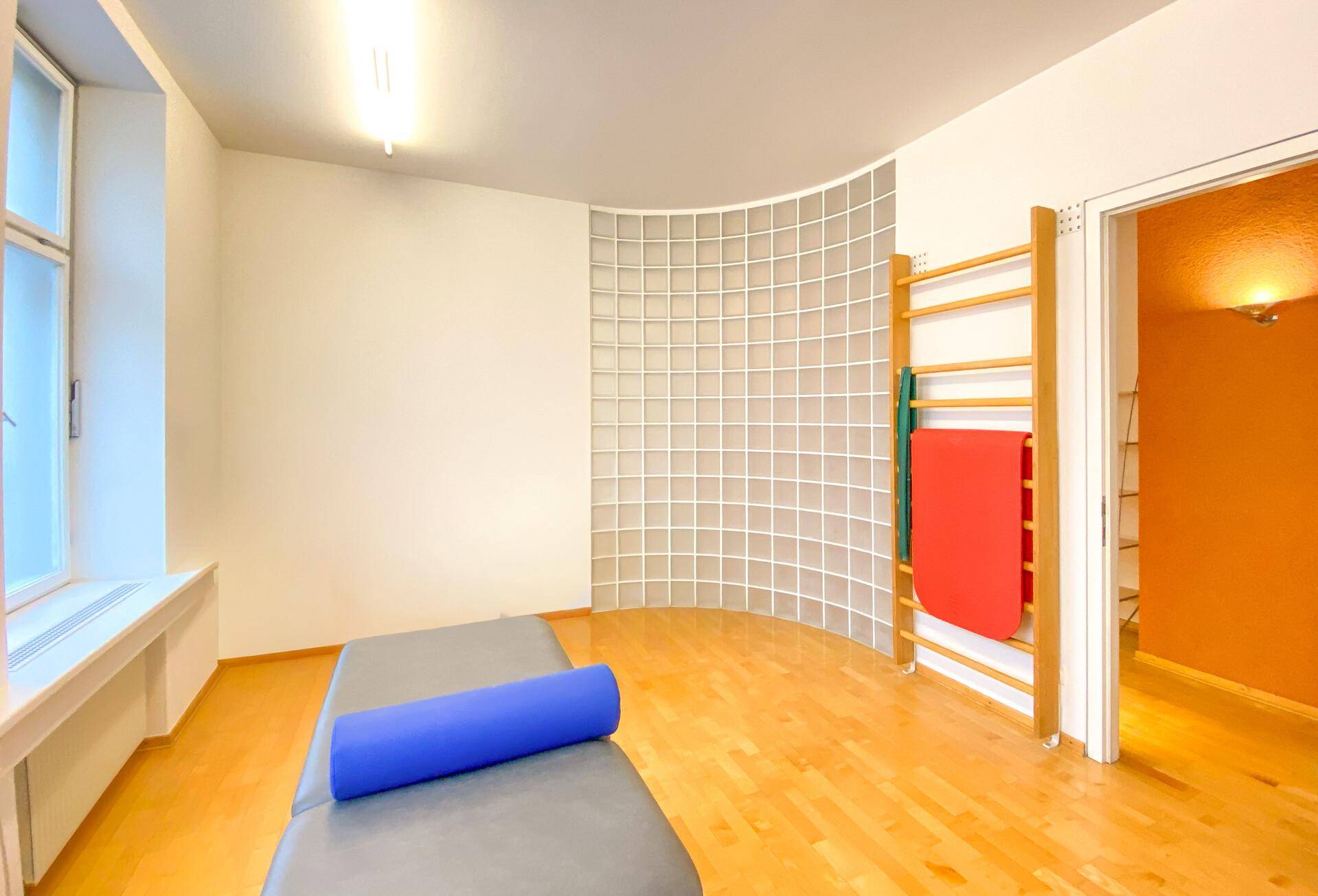 Therapieraum mit Behandlungsliege, Sprossenwand und Glasbausteinwand.