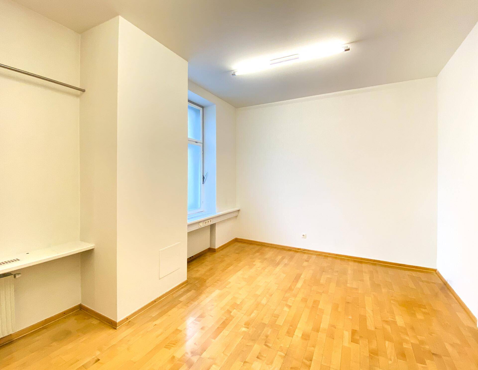 Leerer Büroraum mit Holzboden, Fenster und einfacher Ablagefläche.