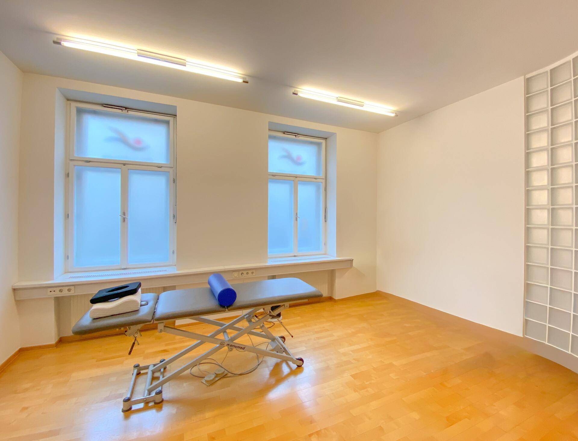 Heller Therapieraum mit zwei Fenstern, Behandlungsliege und effizienter Deckenbeleuchtung.