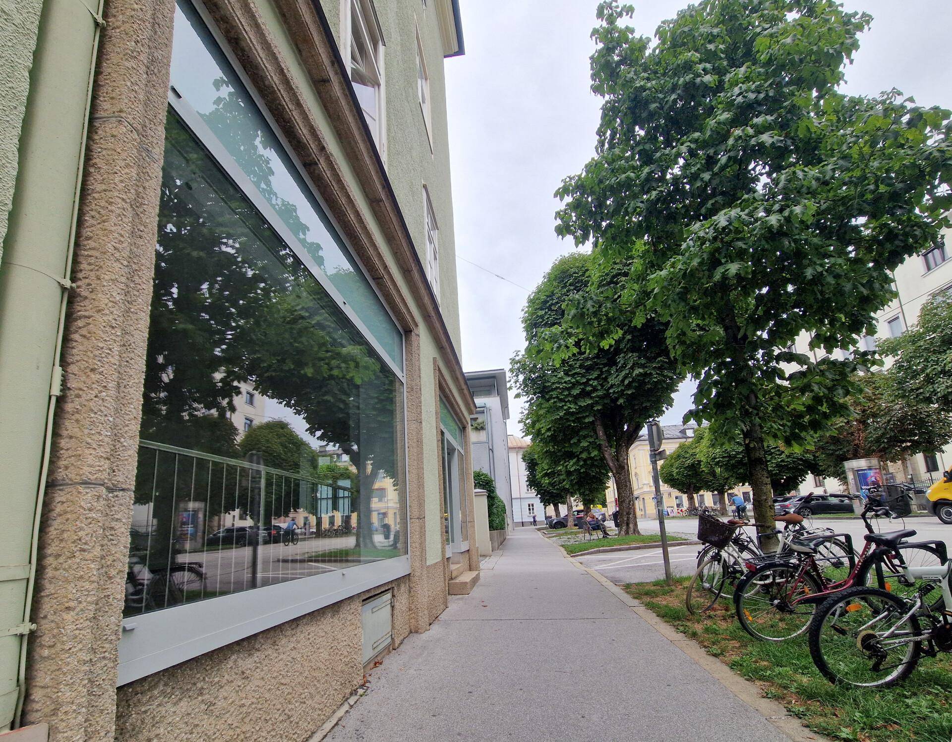 Nahaufnahme der Gebäudefassade im Erdgeschoss mit großen Fenstern, die Bäume und den Gehweg spiegeln.