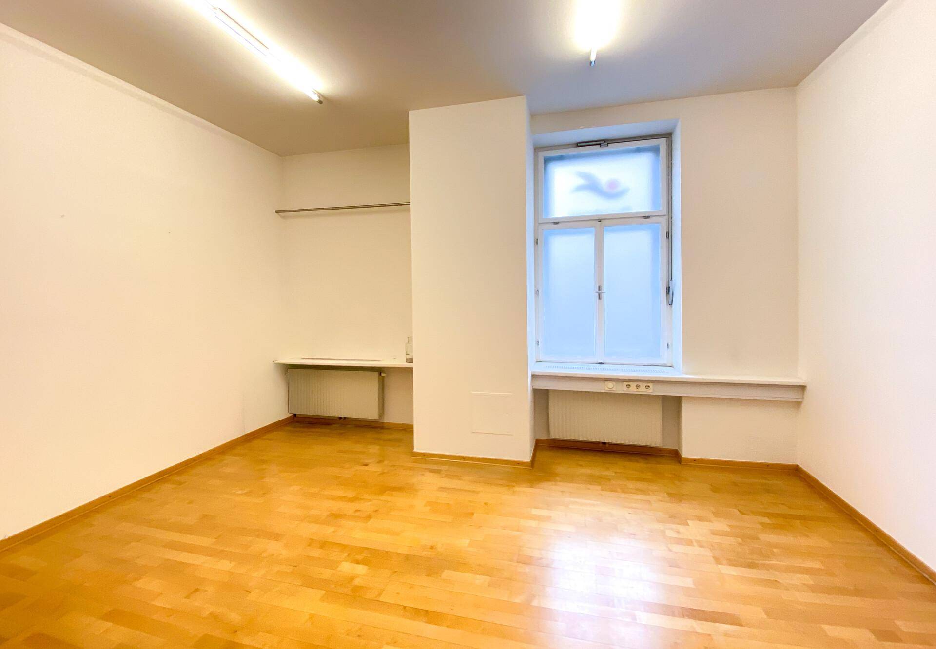 Geräumiger Büroraum mit Holzboden, großem Fenster und schlichter Einrichtung.