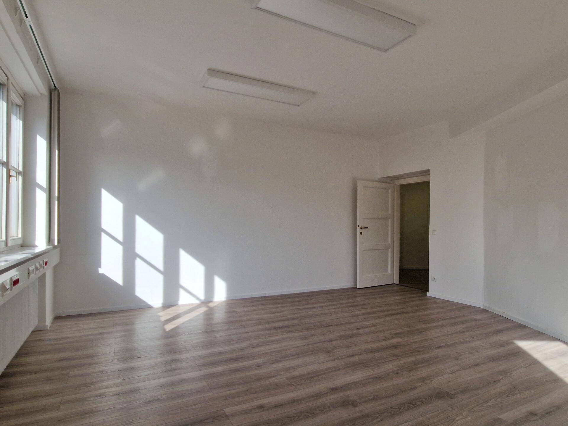 Leerer Büroraum mit hellem Laminatboden, weißen Wänden und einer offenen Tür zu einem weiteren Bereich.