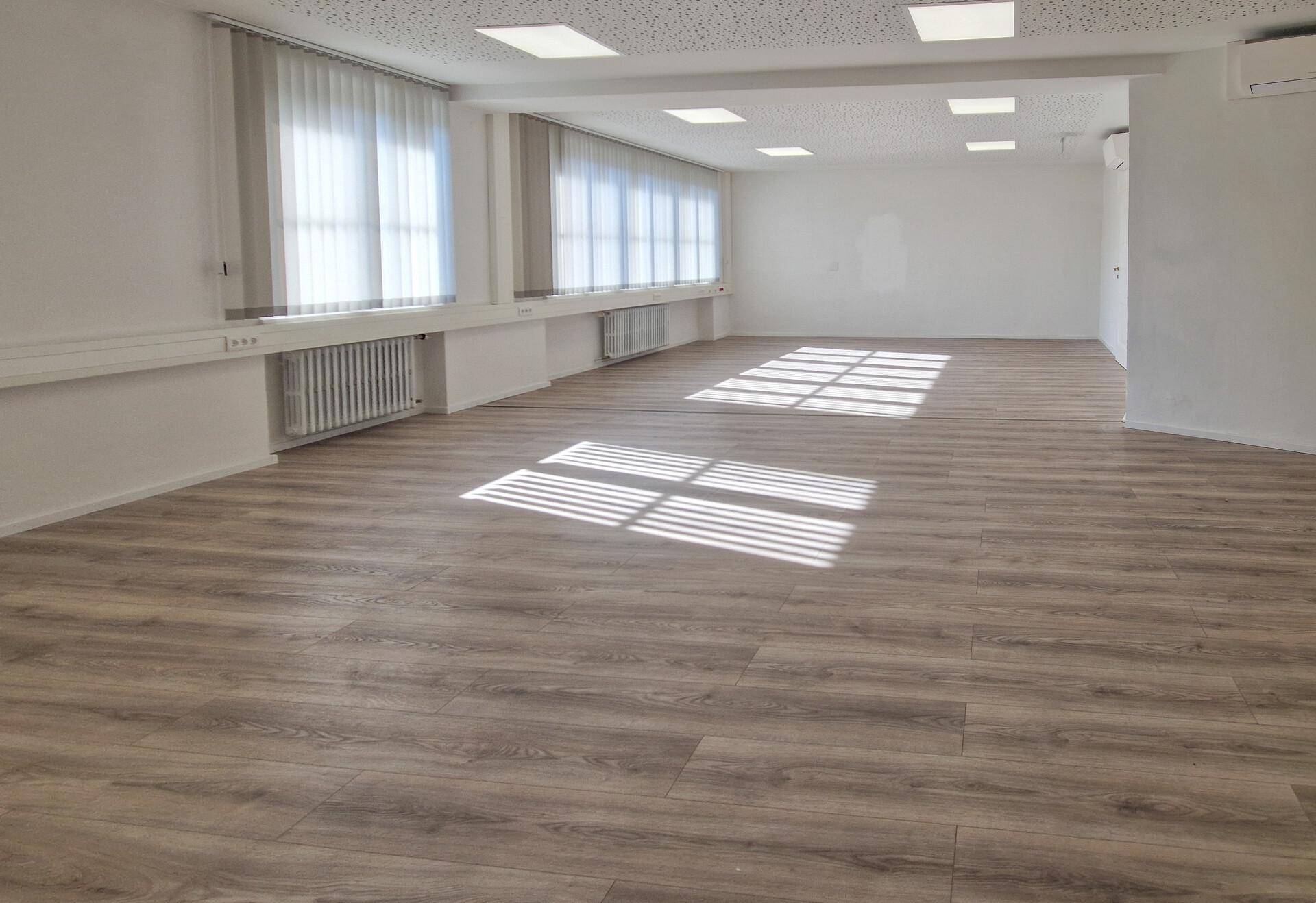 Heller Büroraum mit großen Fenstern, Jalousien und modernem Laminatboden, durchflutet von Sonnenlicht.