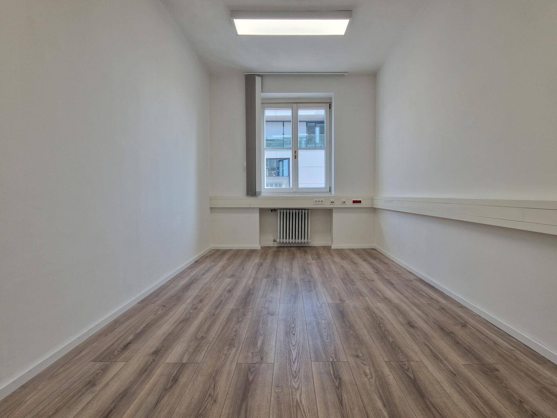 Kleinerer, heller Büroraum mit Laminatboden, weißen Wänden und einem Fenster mit Jalousien.