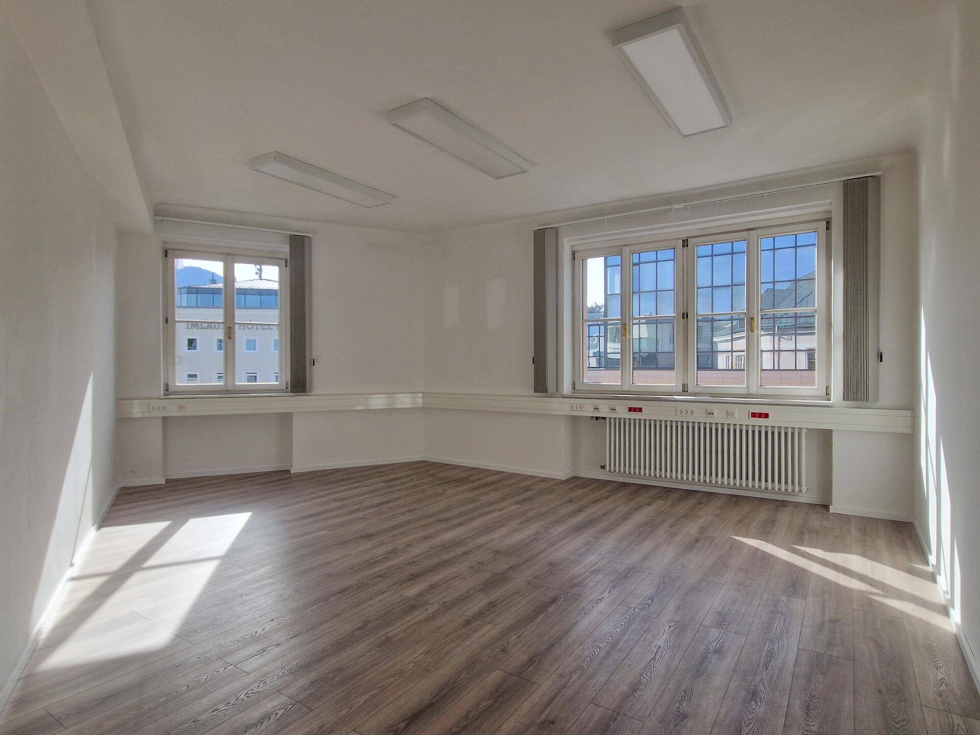 Heller Büroraum mit Laminatboden, weißen Wänden und großen Fenstern, die viel Tageslicht hereinlassen.