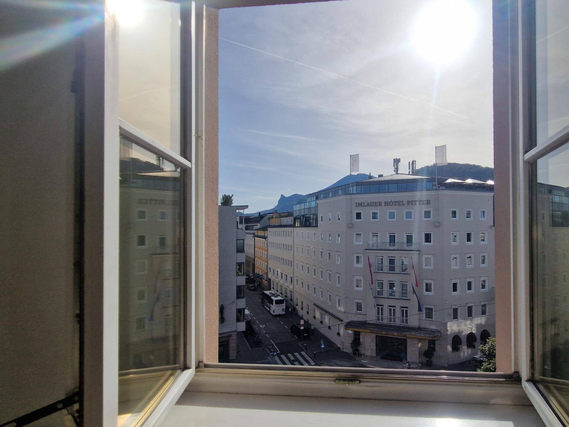 Blick aus einem geöffneten Fenster auf eine belebte Stadtstraße mit Gebäuden und Bergen im Hintergrund.