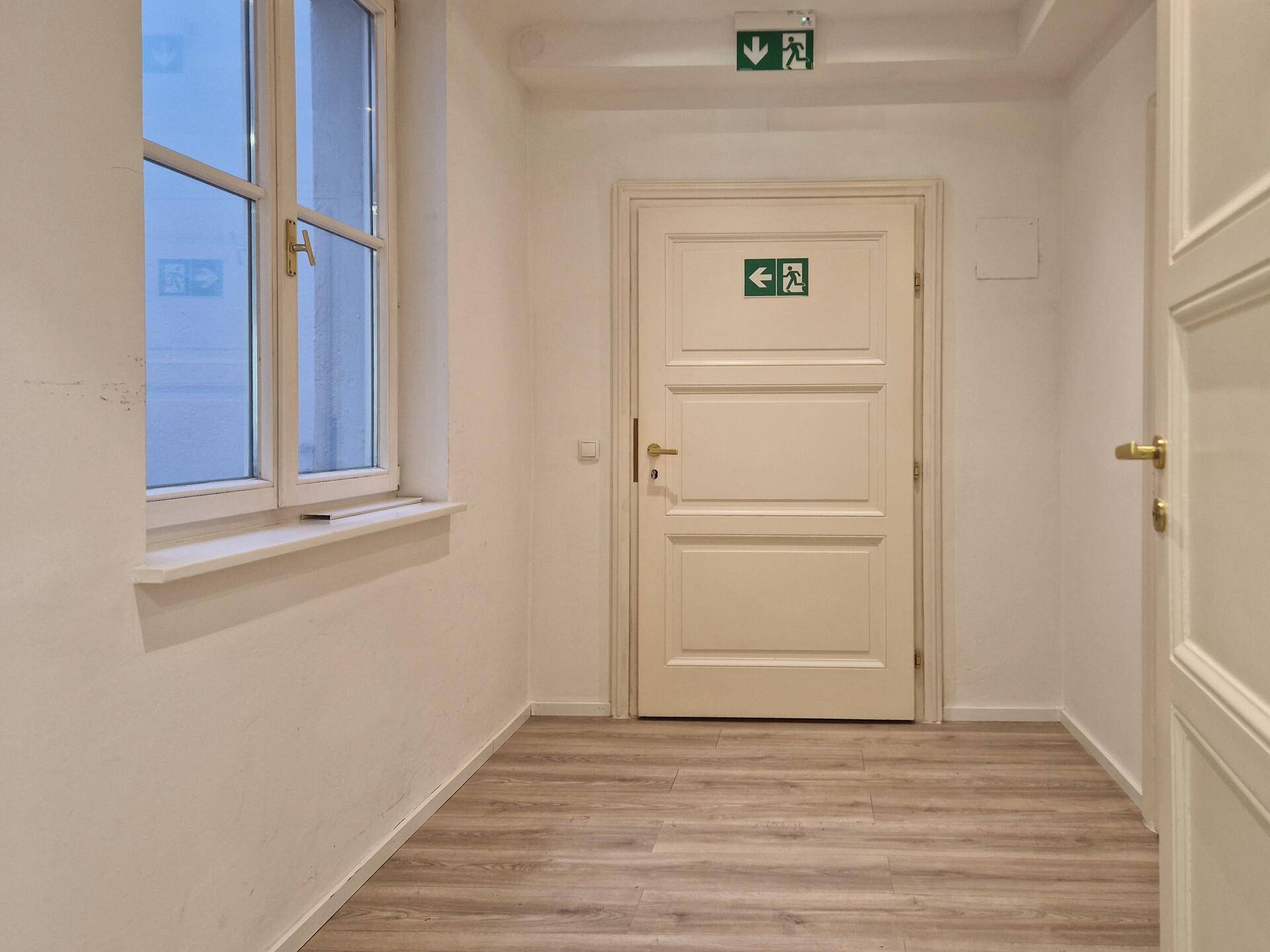 Heller Flur mit Laminatboden, weißen Wänden und einer Tür mit Notausgangsbeschilderung.