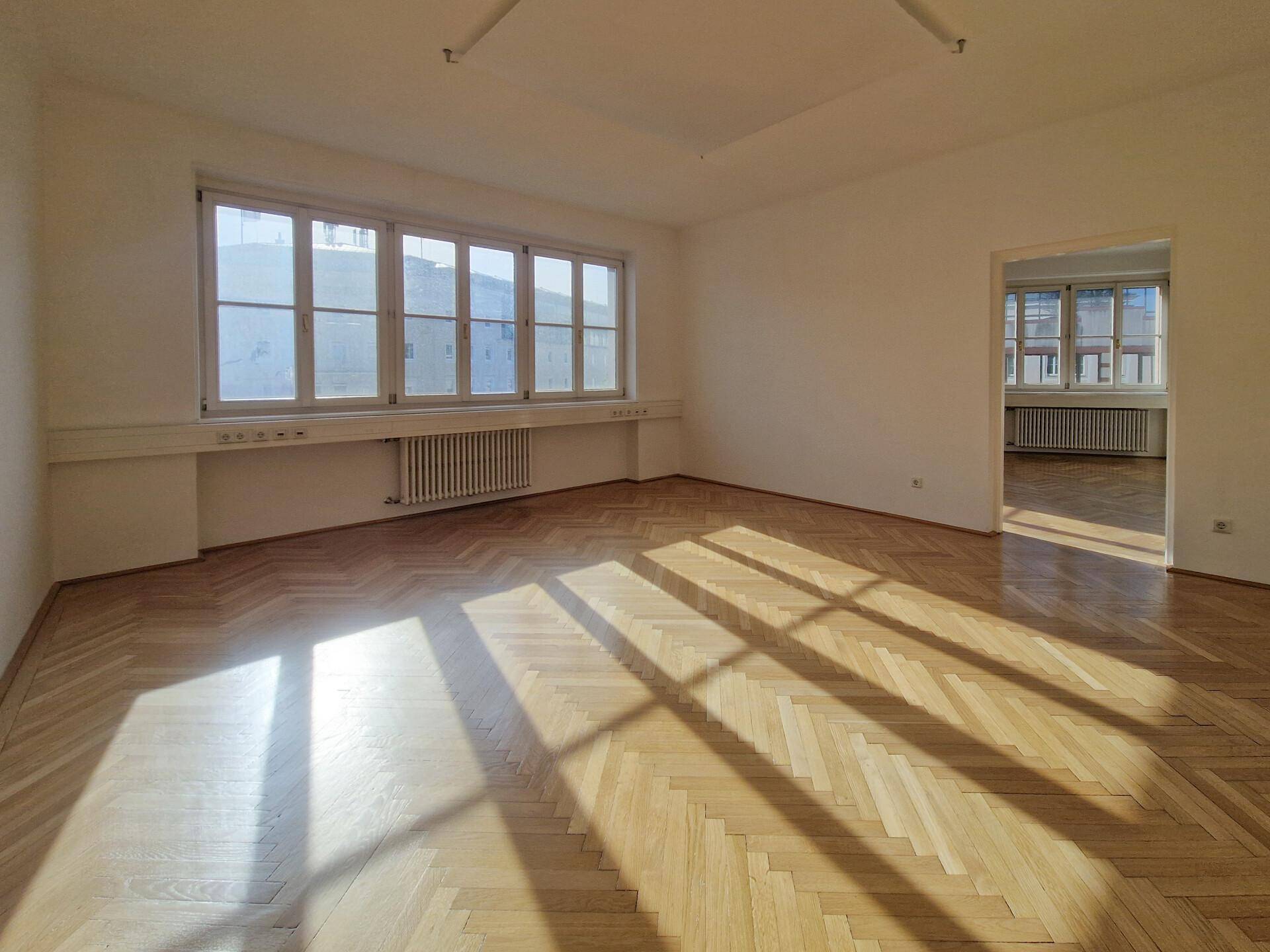 Geräumiger, heller Raum mit großen Fenstern und Fischgrätparkett, durchflutet von Sonnenlicht.