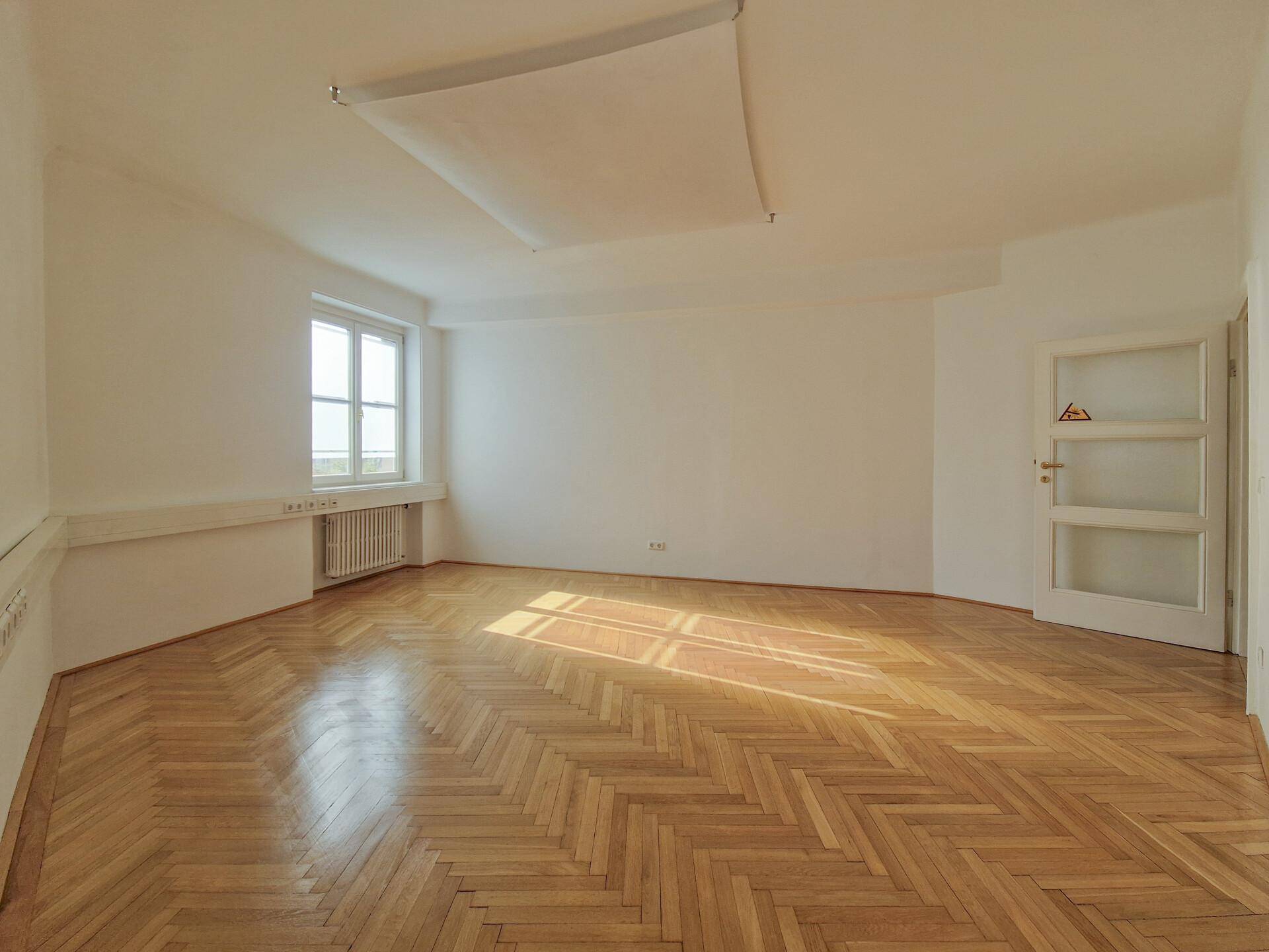 Leerer, heller Raum mit Fischgrätparkett und einem Fenster, das Tageslicht hereinlässt.