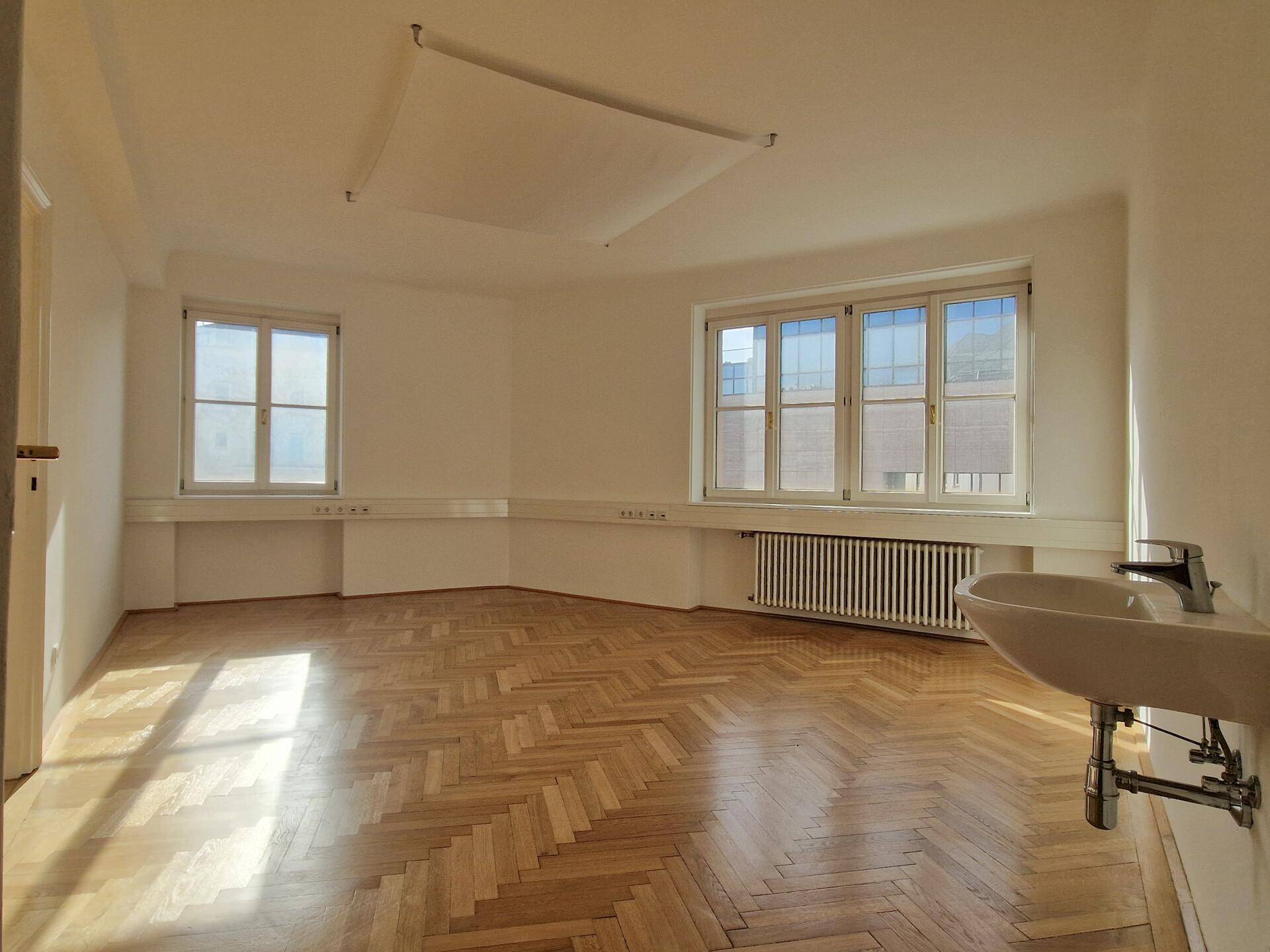 Geräumiger Raum mit Fischgrätparkett, zwei Fenstern und einem Waschbecken an der Wand.