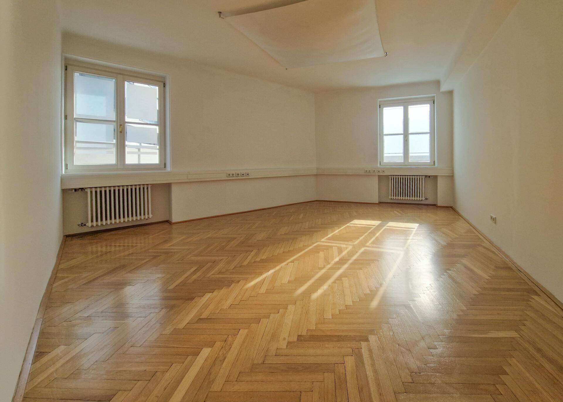 Großer, heller Raum mit Fischgrätparkett und zwei Fenstern, die viel Tageslicht hereinlassen.
