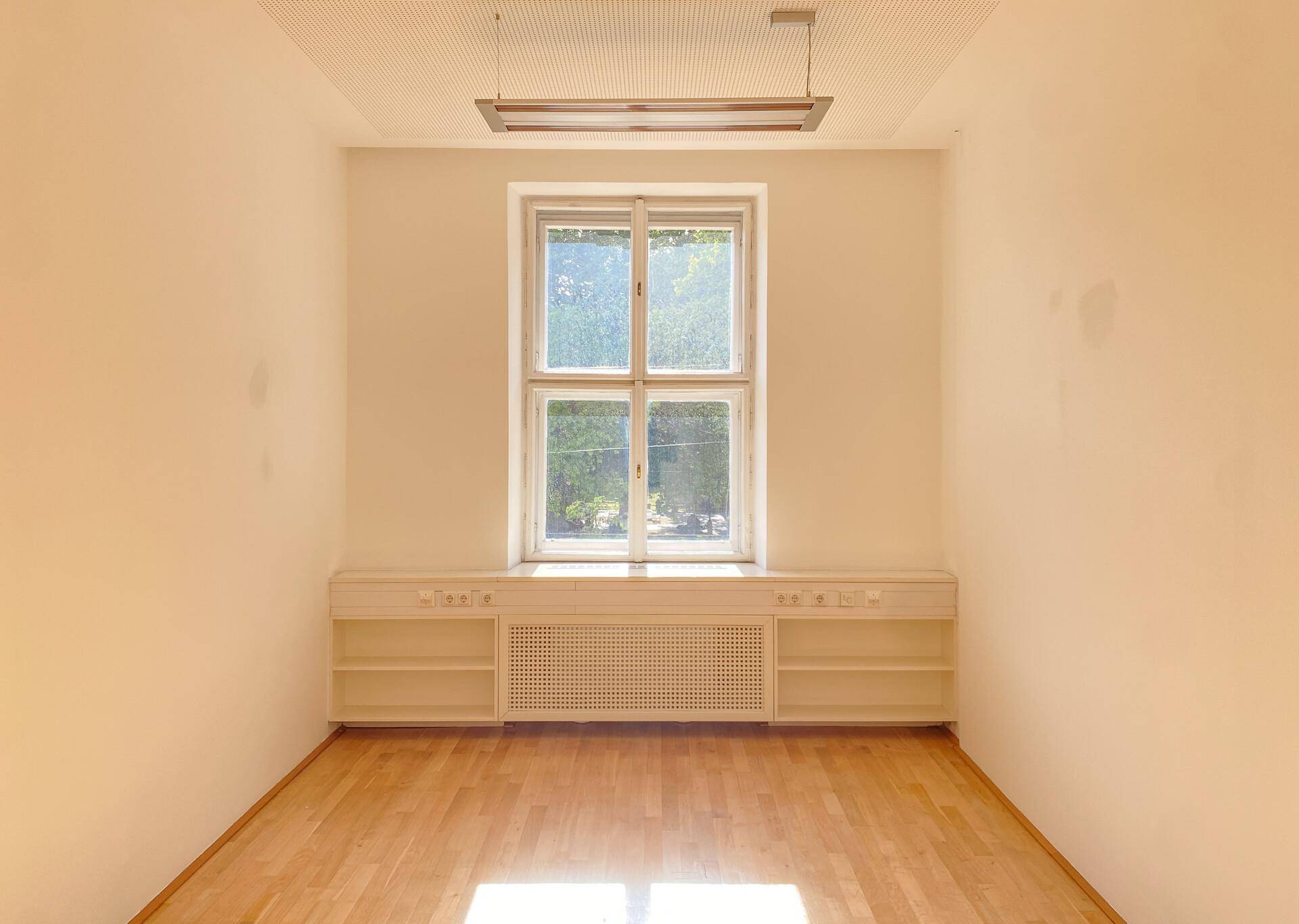 Helles Einzelbüro mit einem großen Fenster, Holzboden und integrierten Heizkörperverkleidungen.