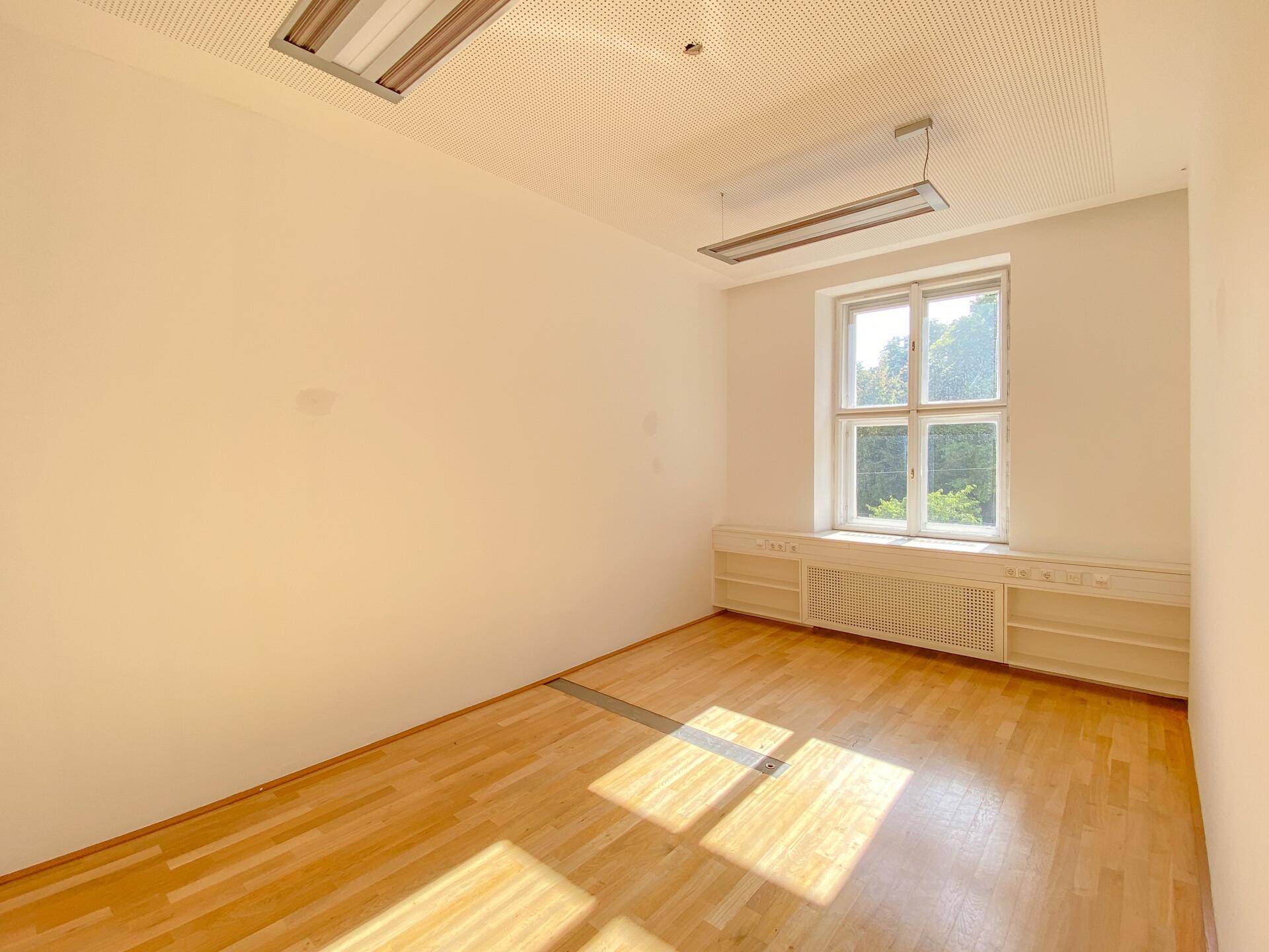 Einzelbüro mit Holzboden, einem großen Fenster und eingebauten Regalen unter dem Fenster.