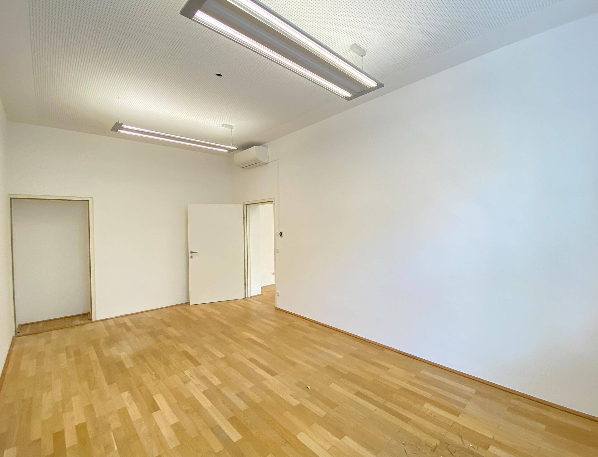 Leerer Büroraum mit Holzboden, weißen Wänden und zwei Türen zu weiteren Räumen.