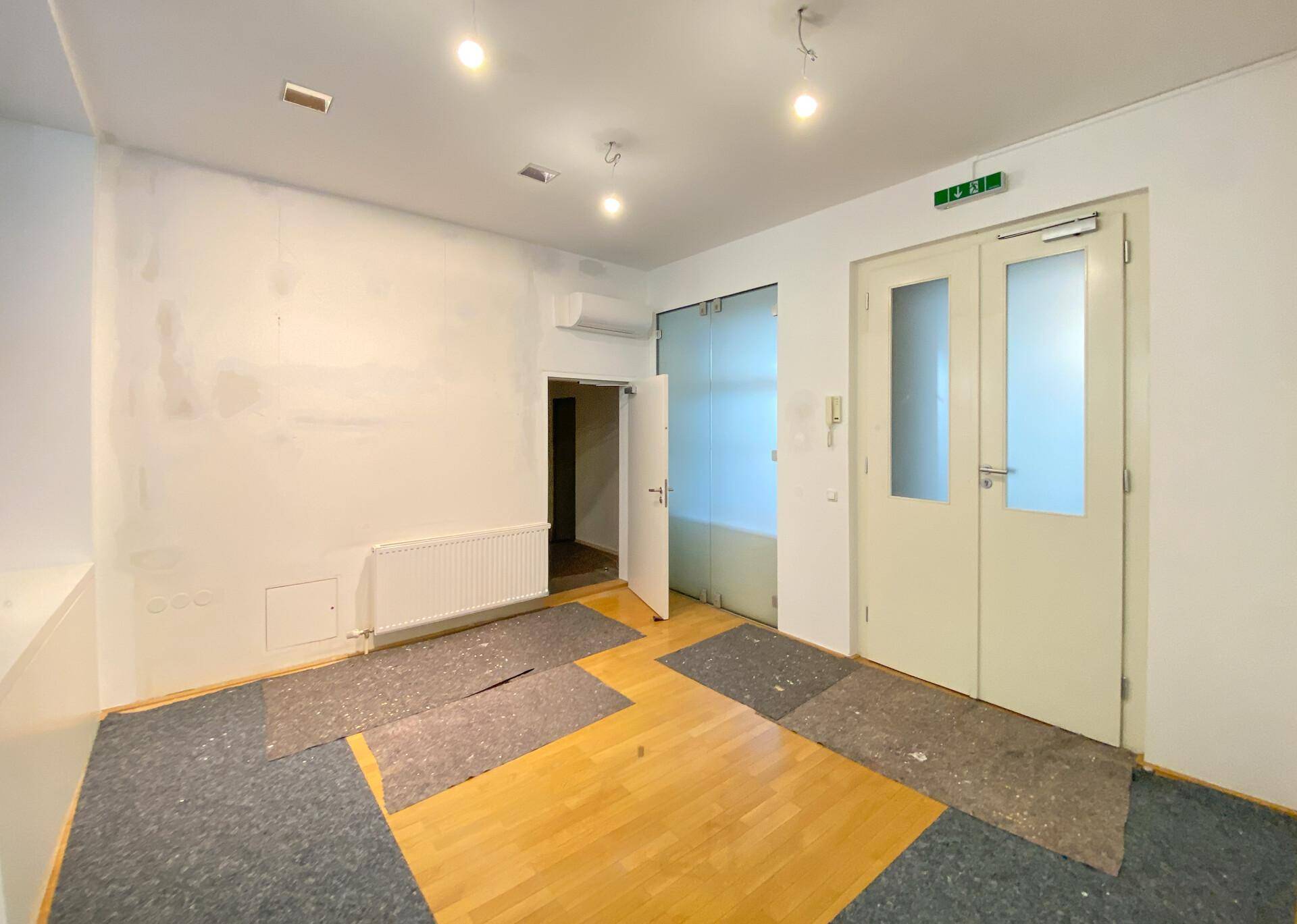 Büroraum mit Holzboden, Teppichresten und zwei großen Eingangstüren mit Glasausschnitten.