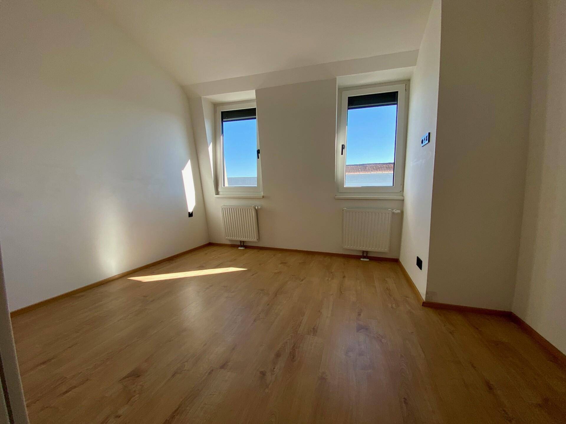 Schlafzimmer mit hellem Holzfußboden und zwei Fenstern, die den Raum gut belichten.