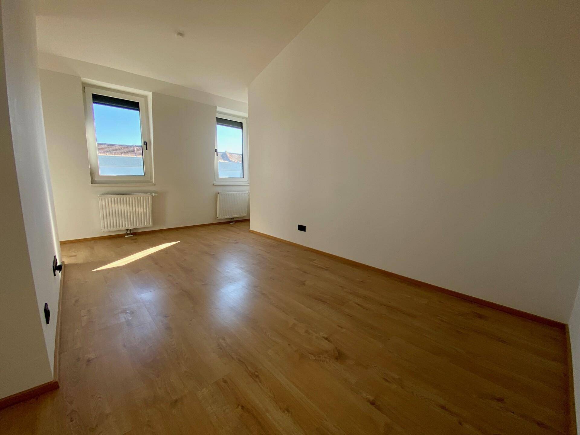 Helles Wohnzimmer mit Holzfußboden und zwei Fenstern, die viel Tageslicht hereinlassen.