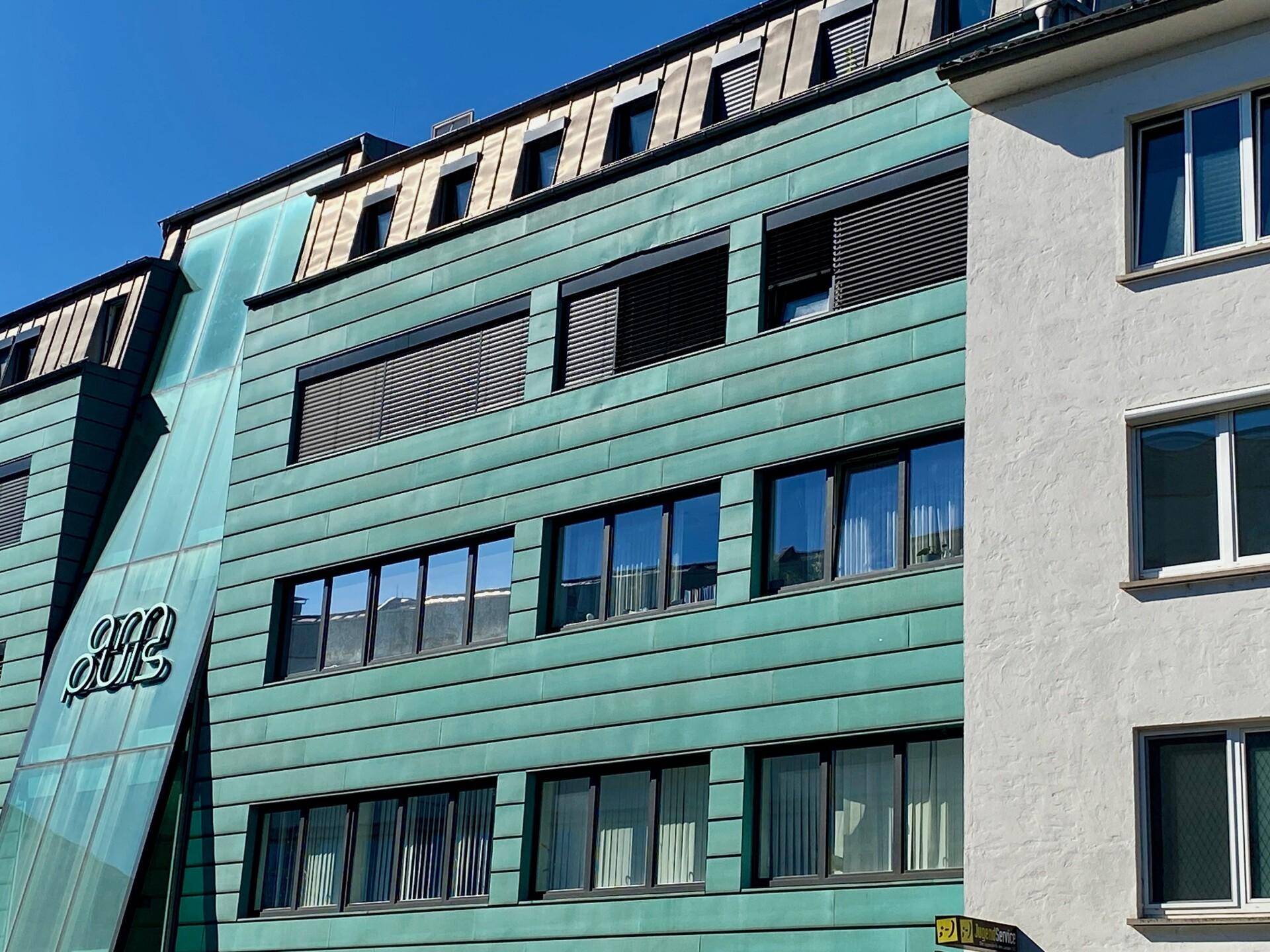 Fassade eines modernen Gebäudes mit grünen Metallpaneelen und zahlreichen Fenstern.