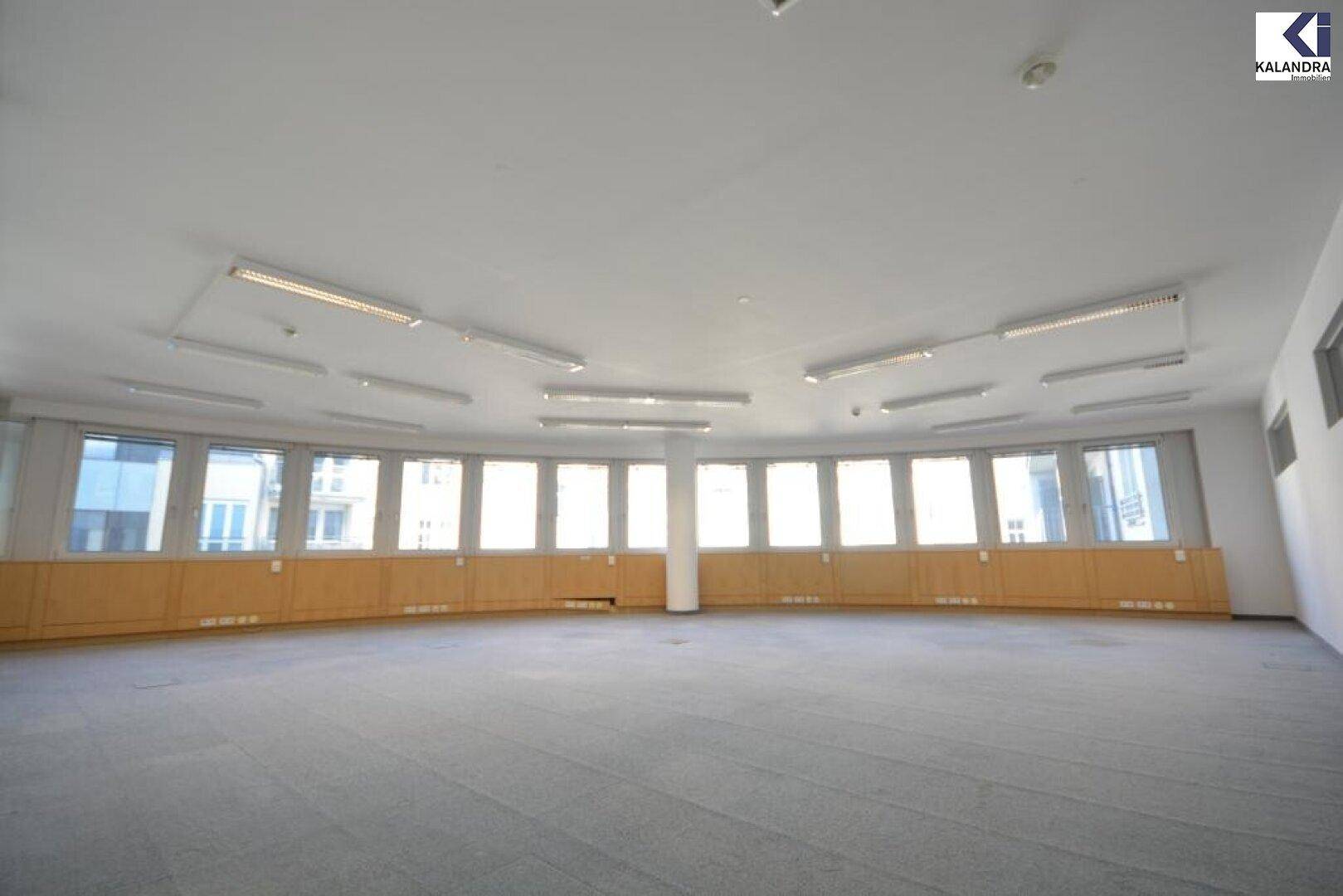 Großes, helles Büro mit Teppichboden und einer langen Reihe von Fenstern, die viel Tageslicht hereinlassen und eine offene Atmosphäre schaffen.