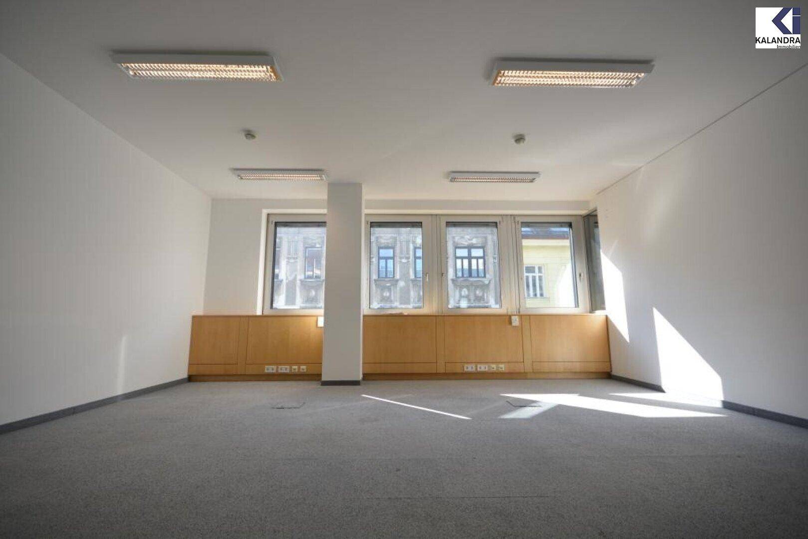 Helles Büro mit Teppichboden, großen Fenstern und Holzverkleidung, gut beleuchtet durch Tageslicht, bietet eine angenehme Arbeitsumgebung.