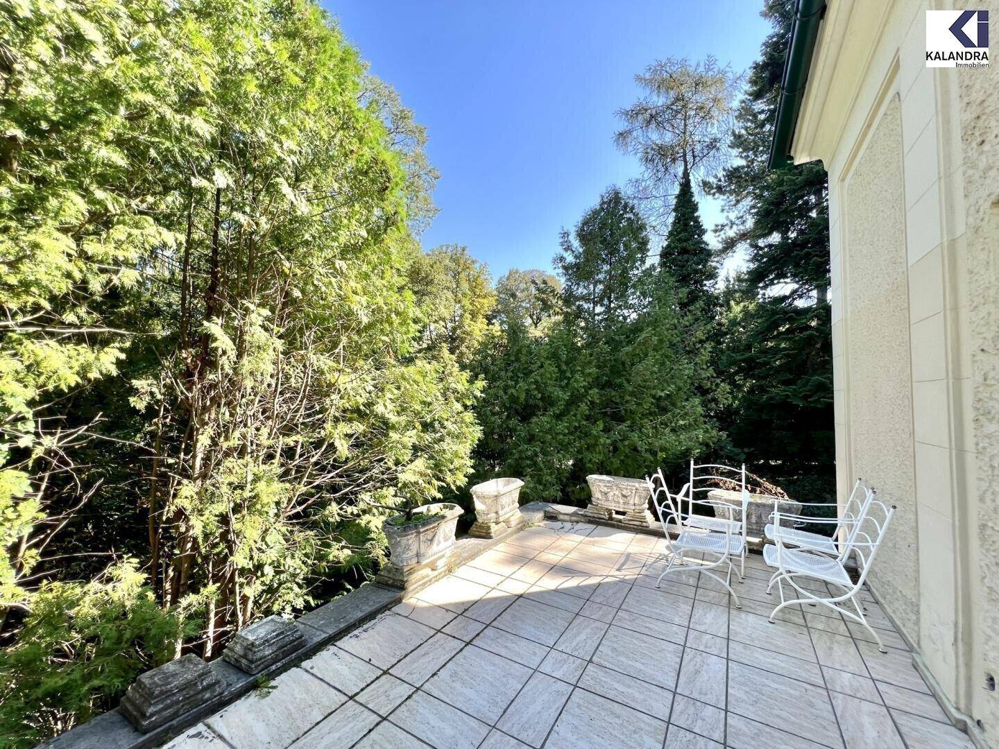 Sonniger Terrassenbereich mit Fliesenboden und Gartenmöbeln, umgeben von üppigem Grün.